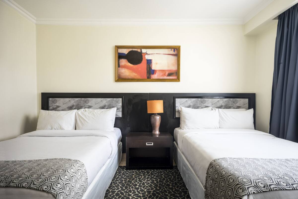 A hotel room with two beds, a nightstand with a lamp, a painting on the wall, and a patterned carpet.
