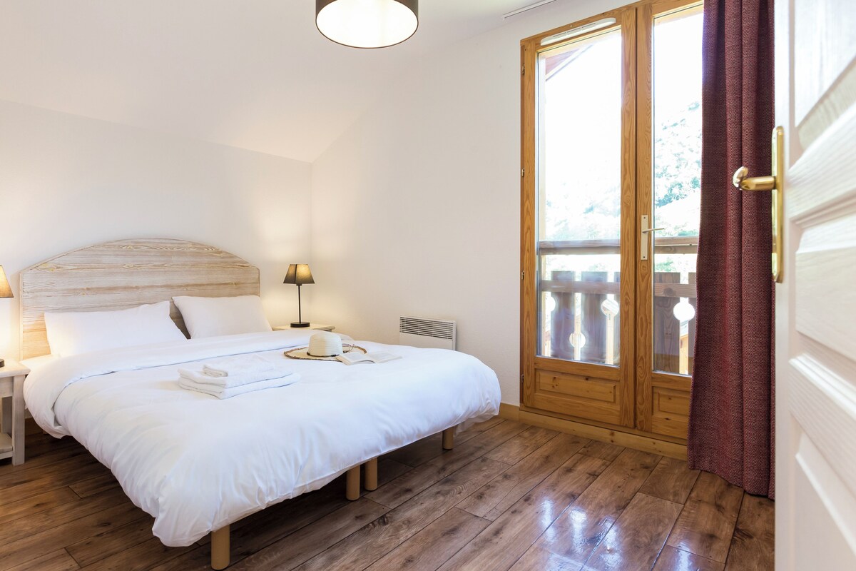 A bedroom with a wooden headboard, a bedside table with a lamp, a large window with curtains, and a wooden floor.