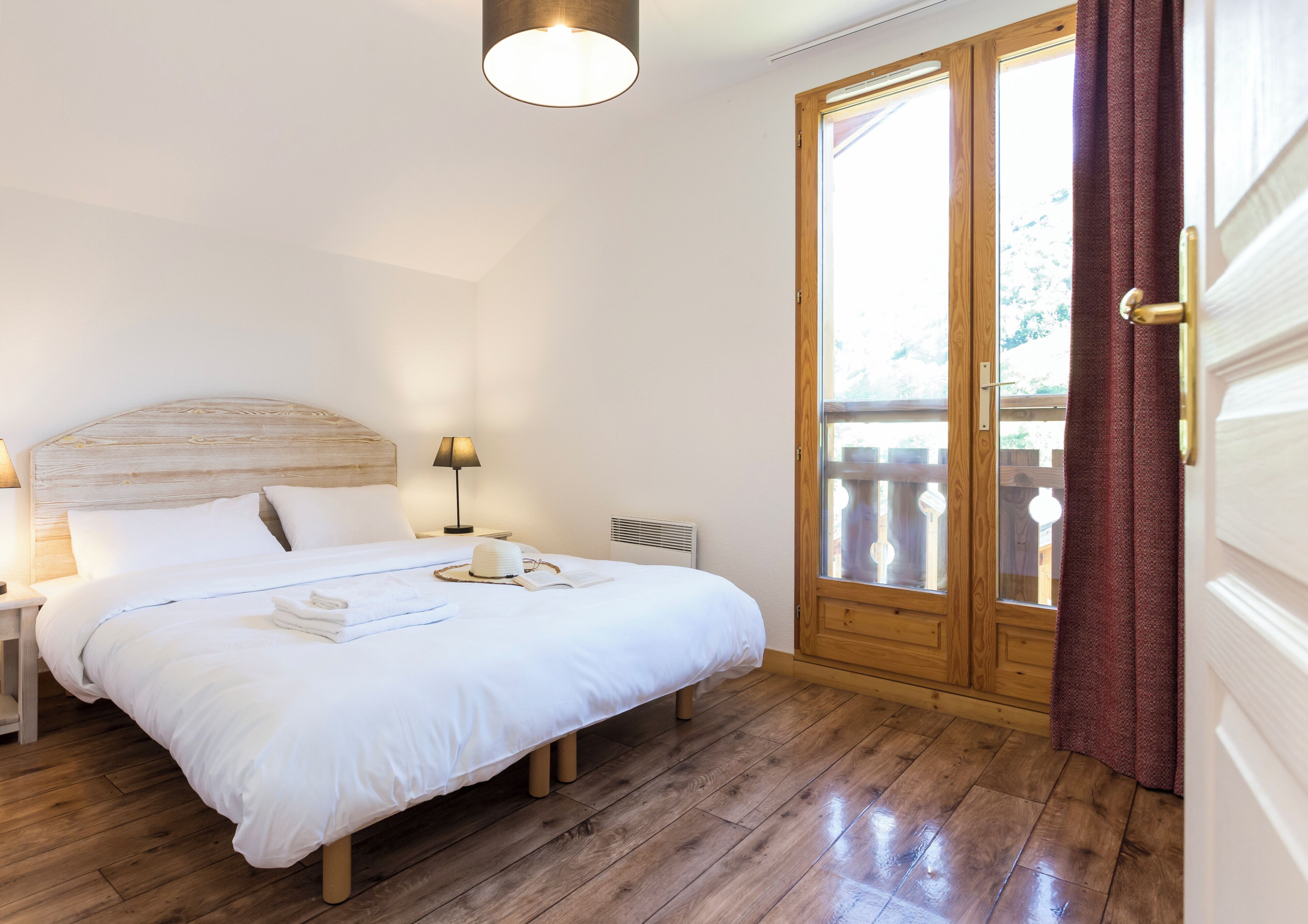 Un dormitorio con cabecera de madera, lámpara de noche, un ventanal con cortinas y piso de madera.