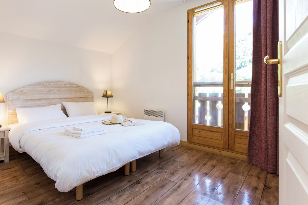 A bedroom with a wooden headboard, a bedside table with a lamp, a large window with curtains, and a wooden floor.