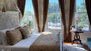 A bedroom with a large bed, a bedside table with a lamp, and a view of the outdoors through multiple windows. - Hoyt House Amelia Island (Fernandina Beach)