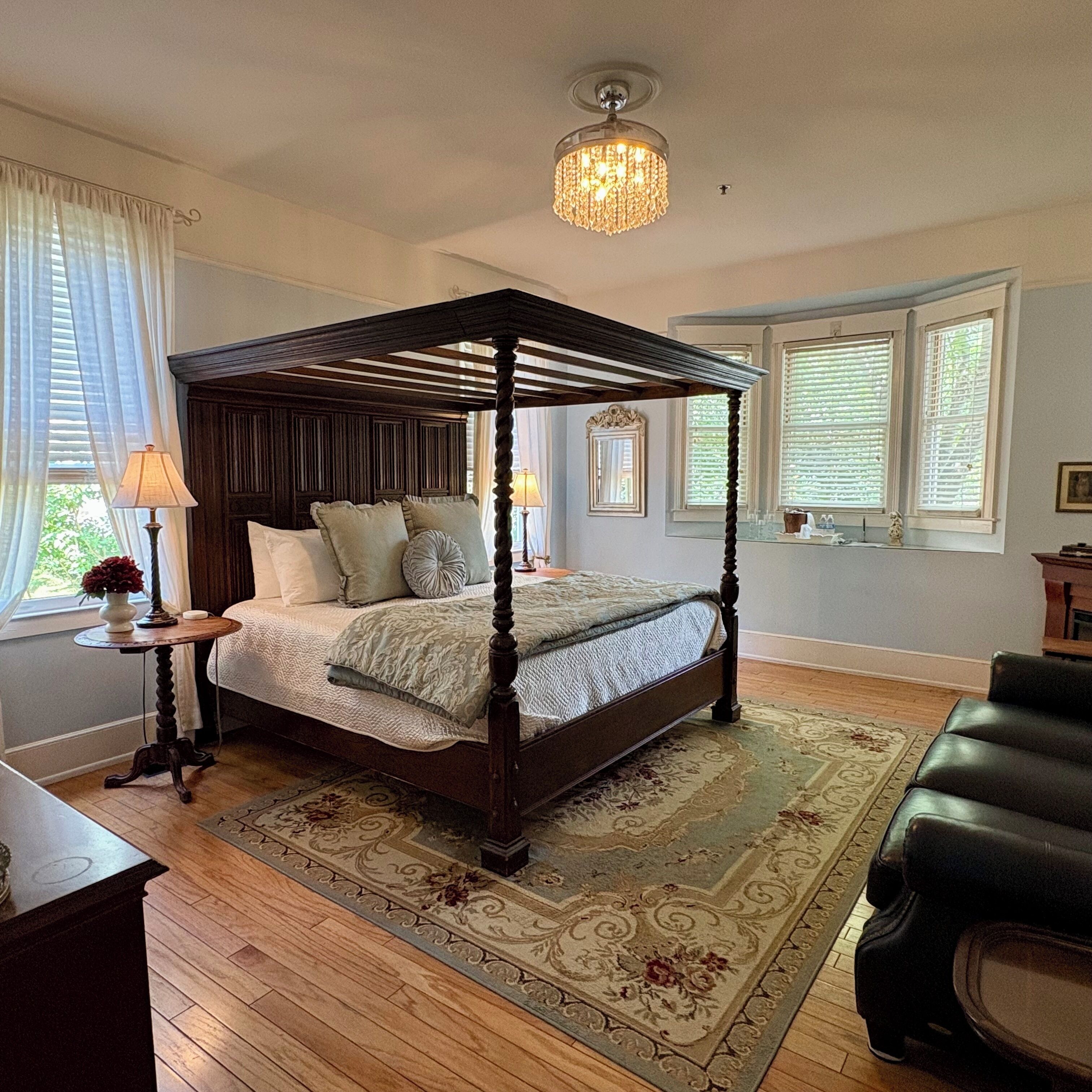 A four-poster bed with a wooden frame, a chandelier above, and a view of the outdoors through the windows.