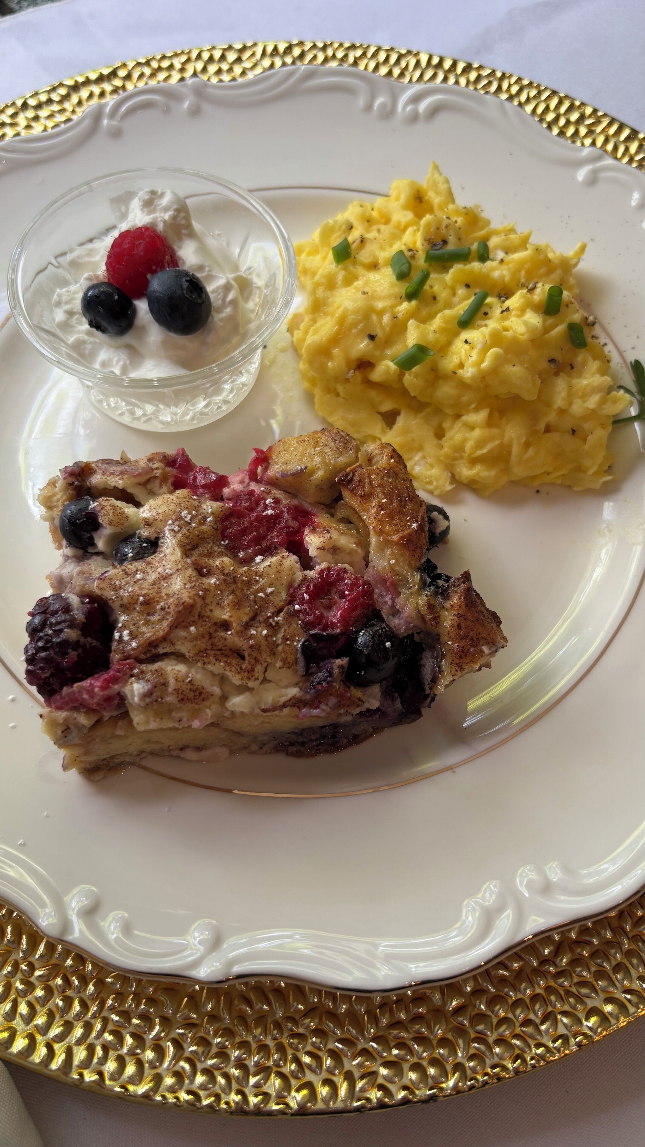 A plate with scrambled eggs, a slice of berry-topped cake, and a small bowl of yogurt with berries.