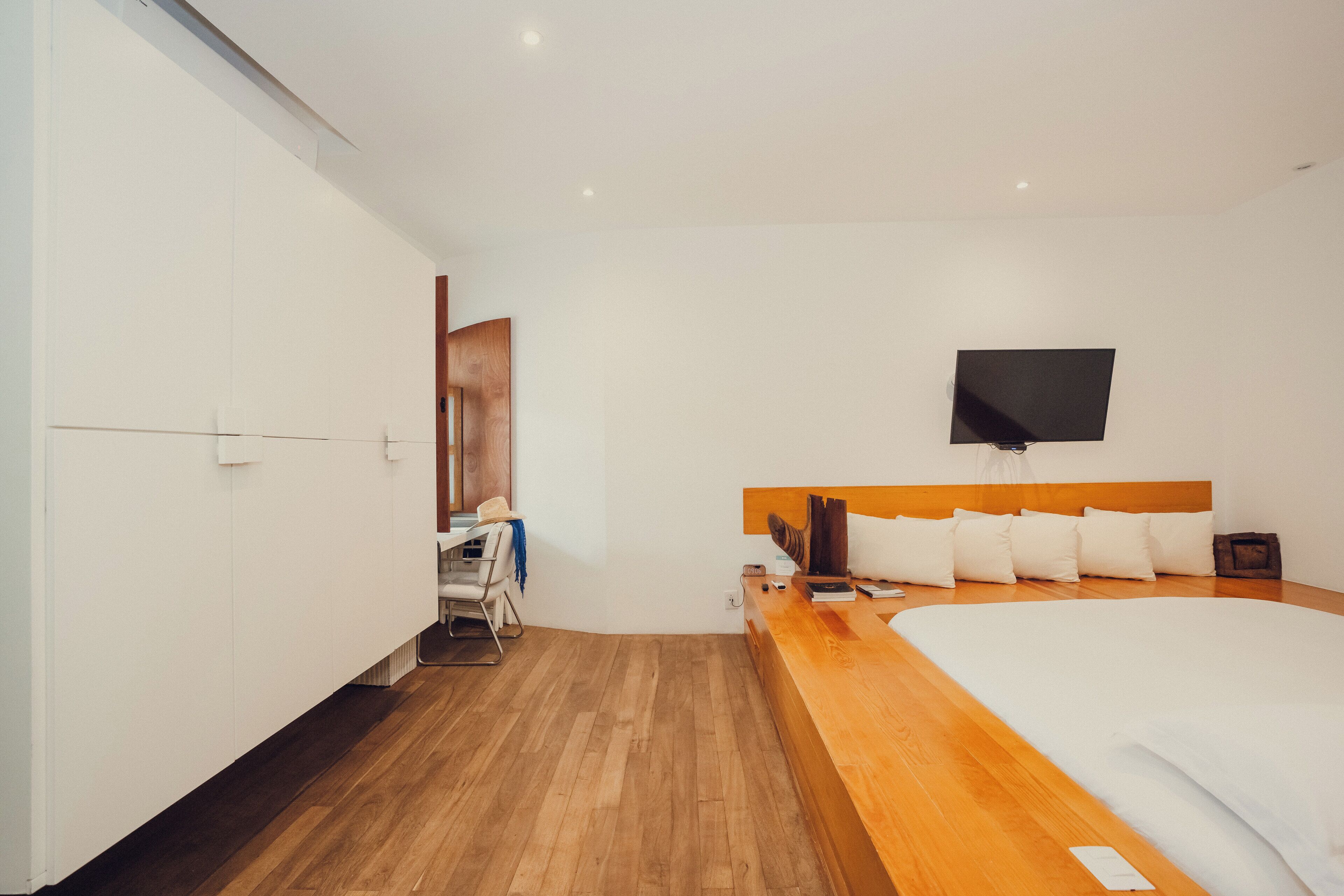 A modern hotel room with a wooden floor, a large bed with white bedding, a flat-screen TV mounted on the wall, and a small desk with a chair.