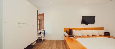 A modern hotel room with a wooden floor, a large bed with white bedding, a flat-screen TV mounted on the wall, and a small desk with a chair.