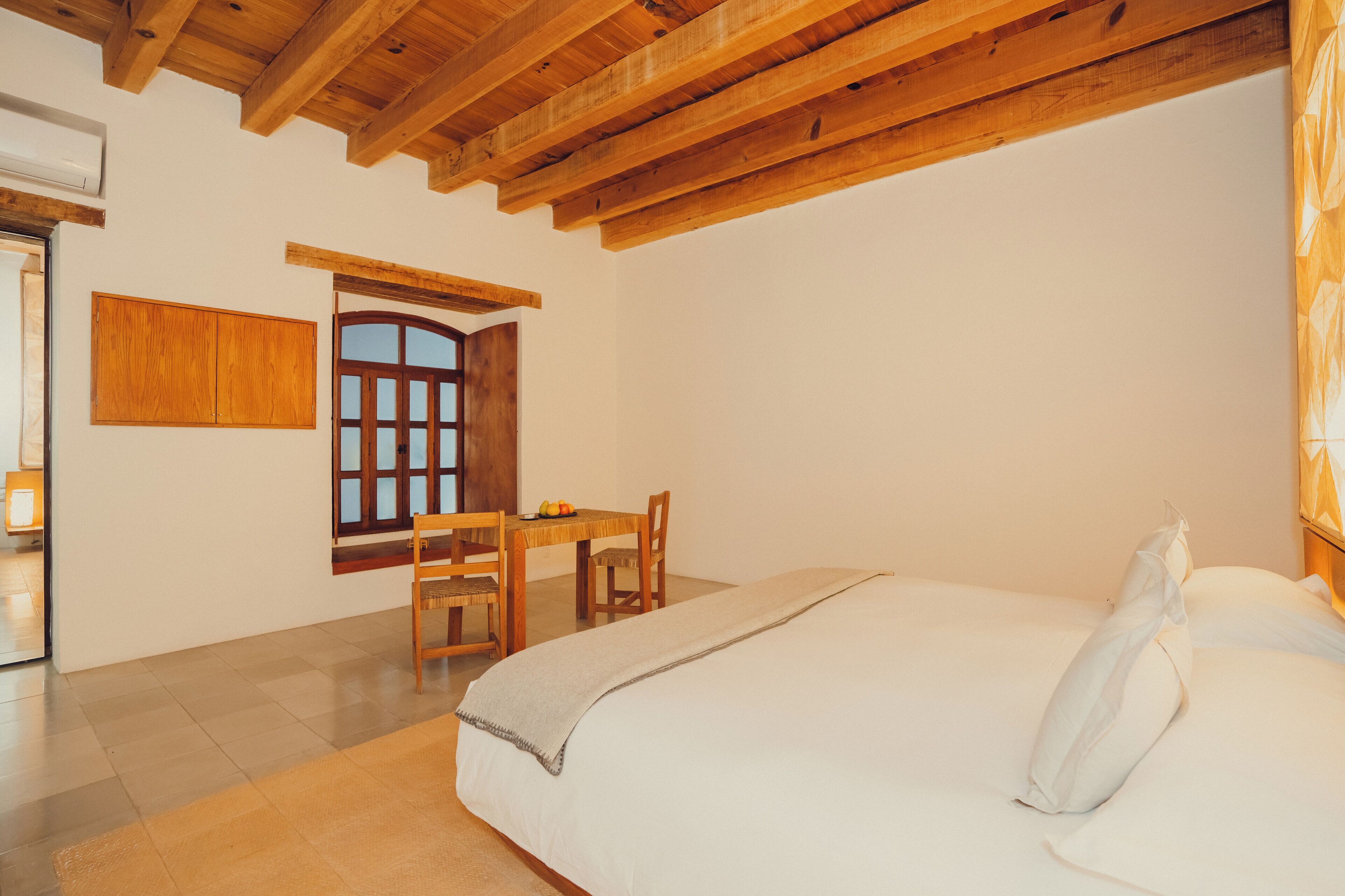 A bedroom with a wooden ceiling, a bed with white linens, a wooden table with chairs, and a window with wooden shutters.
