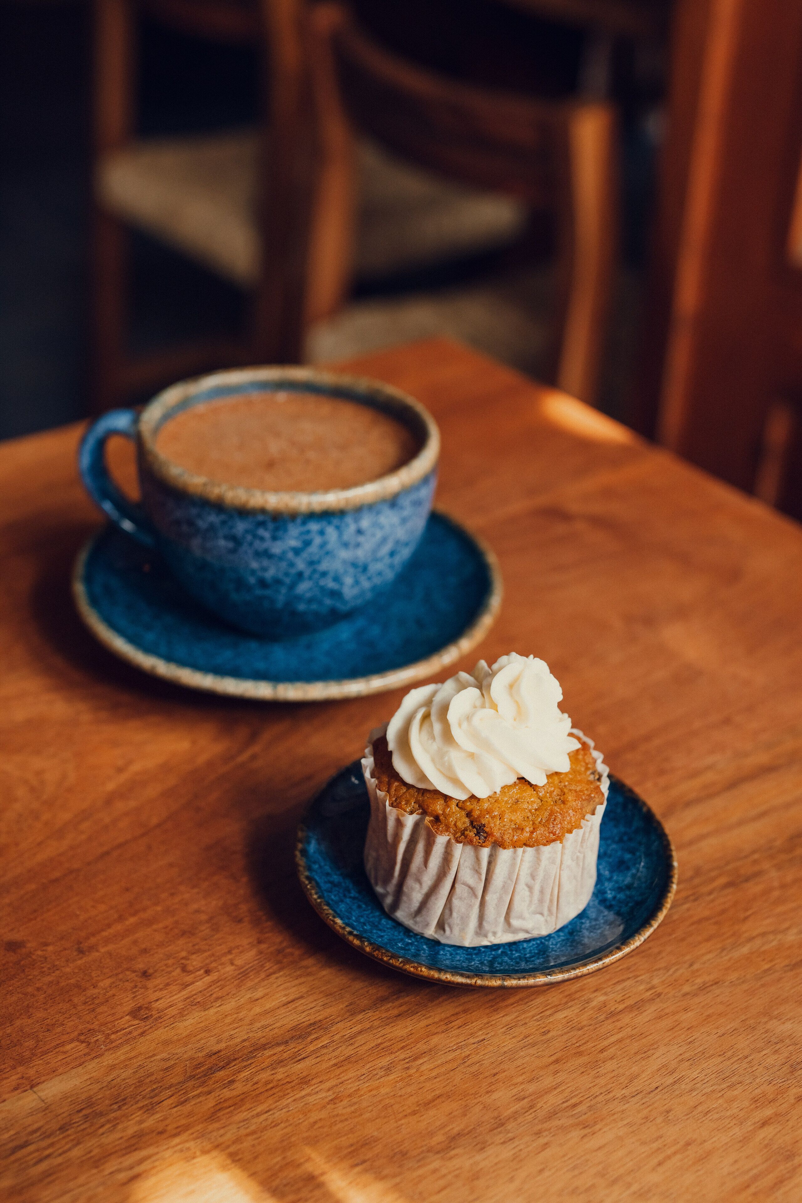 Ein Holztisch mit einer blauen Keramiktasse und einem Untertasse sowie einem Cupcake mit Schlagsahne auf einem passenden blauen Teller.