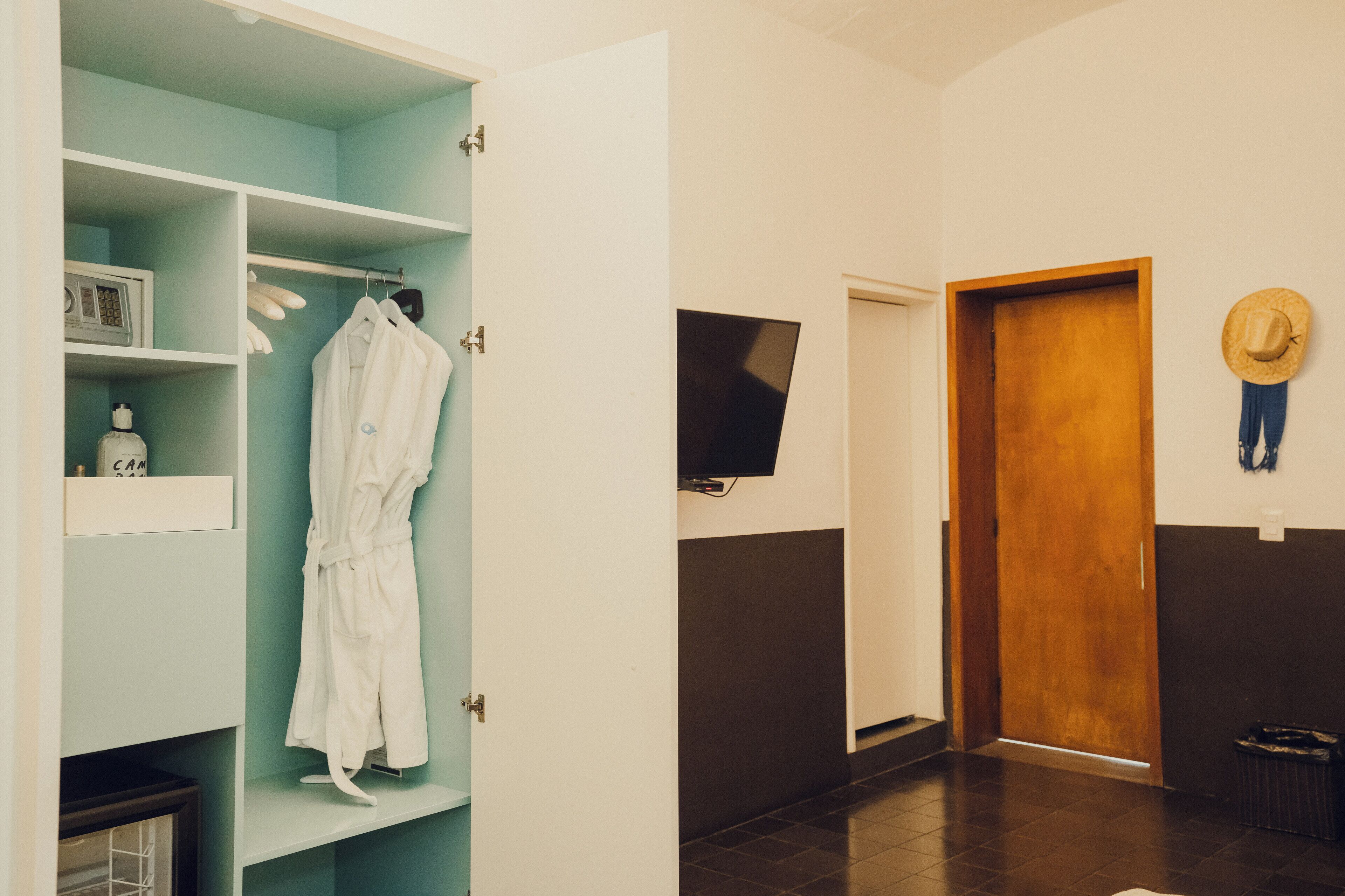 A hotel room with a teal-colored wardrobe, a white robe hanging, a television mounted on the wall, and a wooden door.