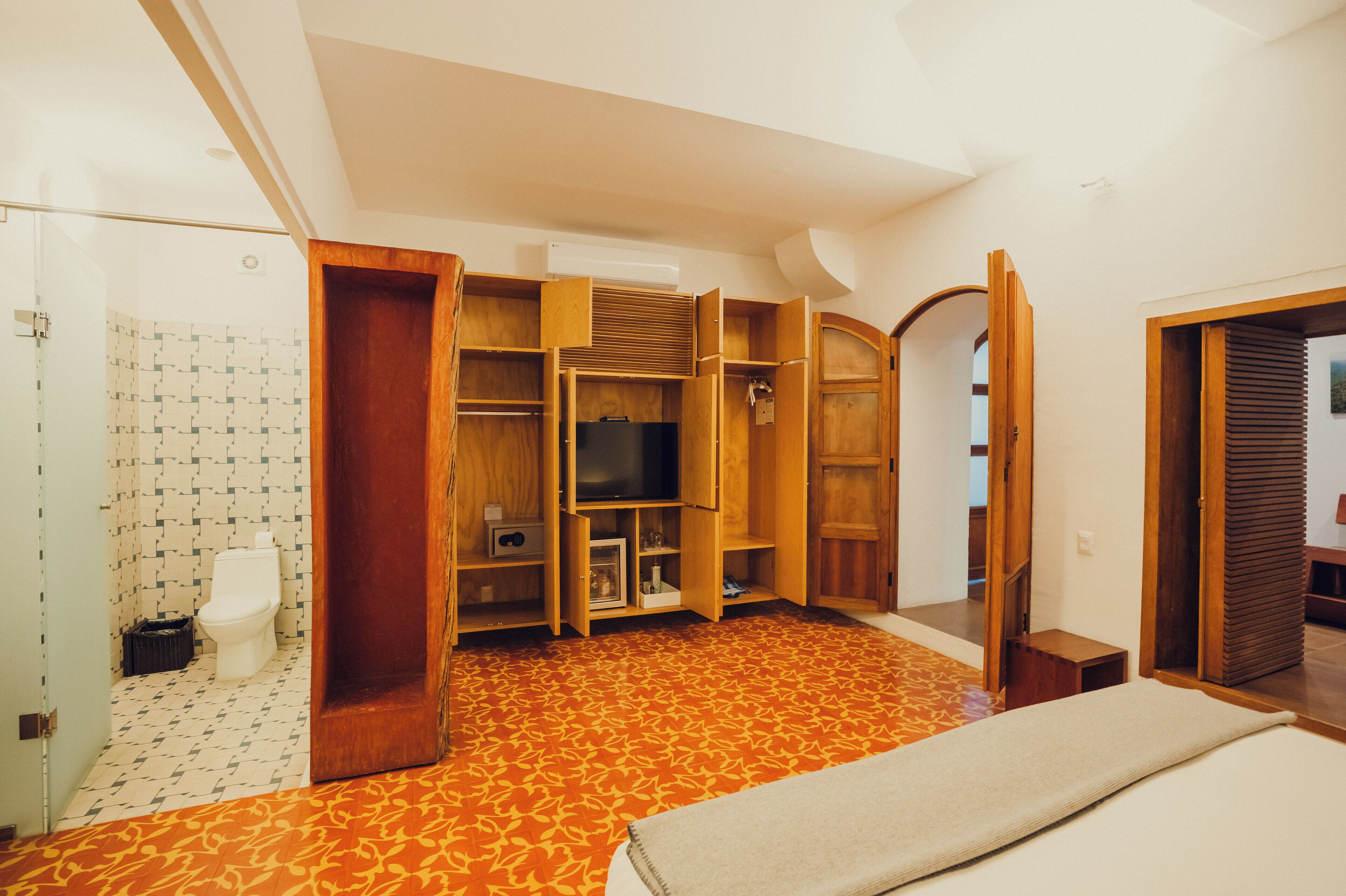 A room with a patterned floor, wooden furniture, a television, and a bathroom with a toilet and patterned tiles.