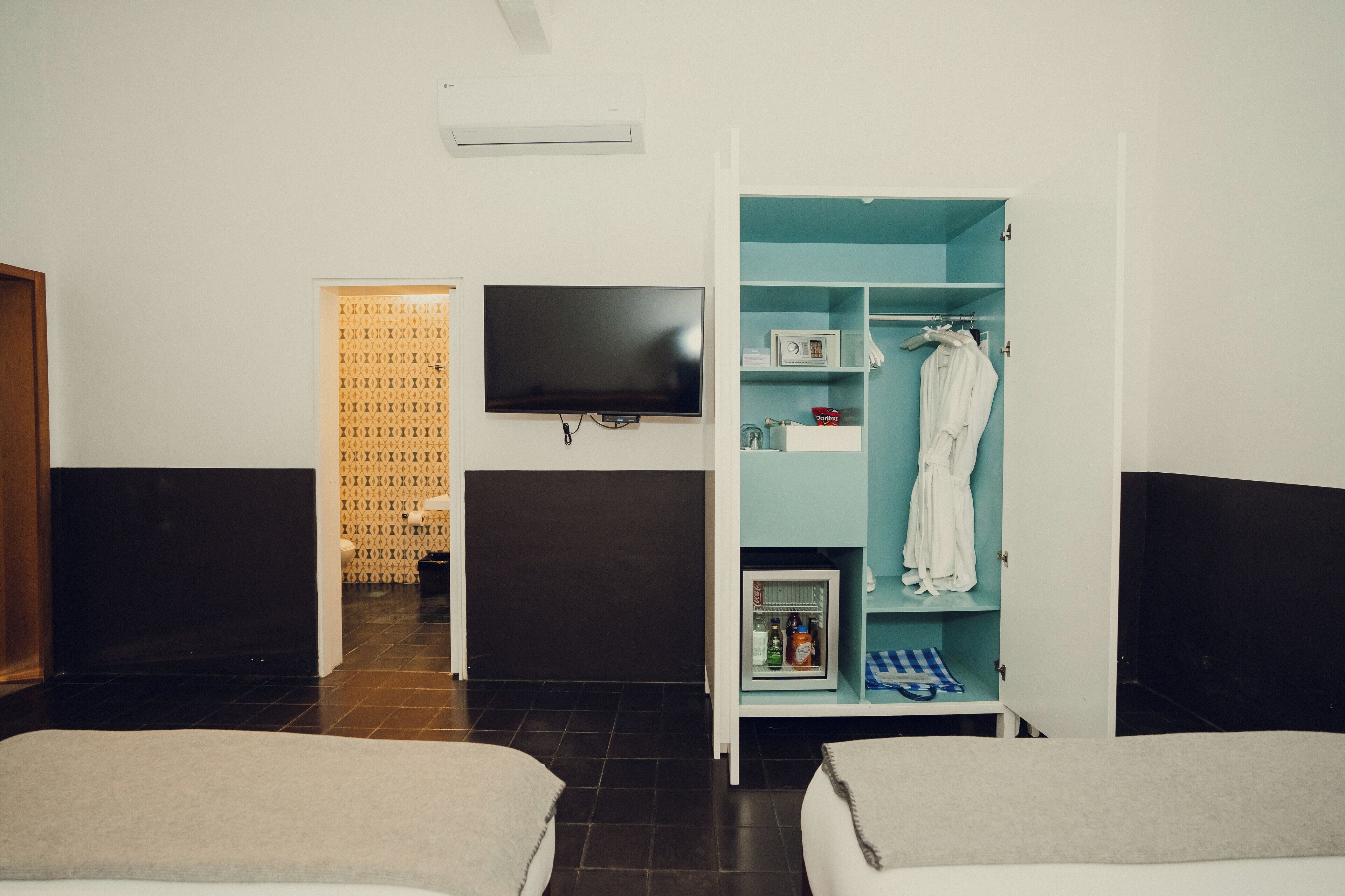 A hotel room with a bed, a TV mounted on the wall, a wardrobe with shelves, and a small refrigerator.