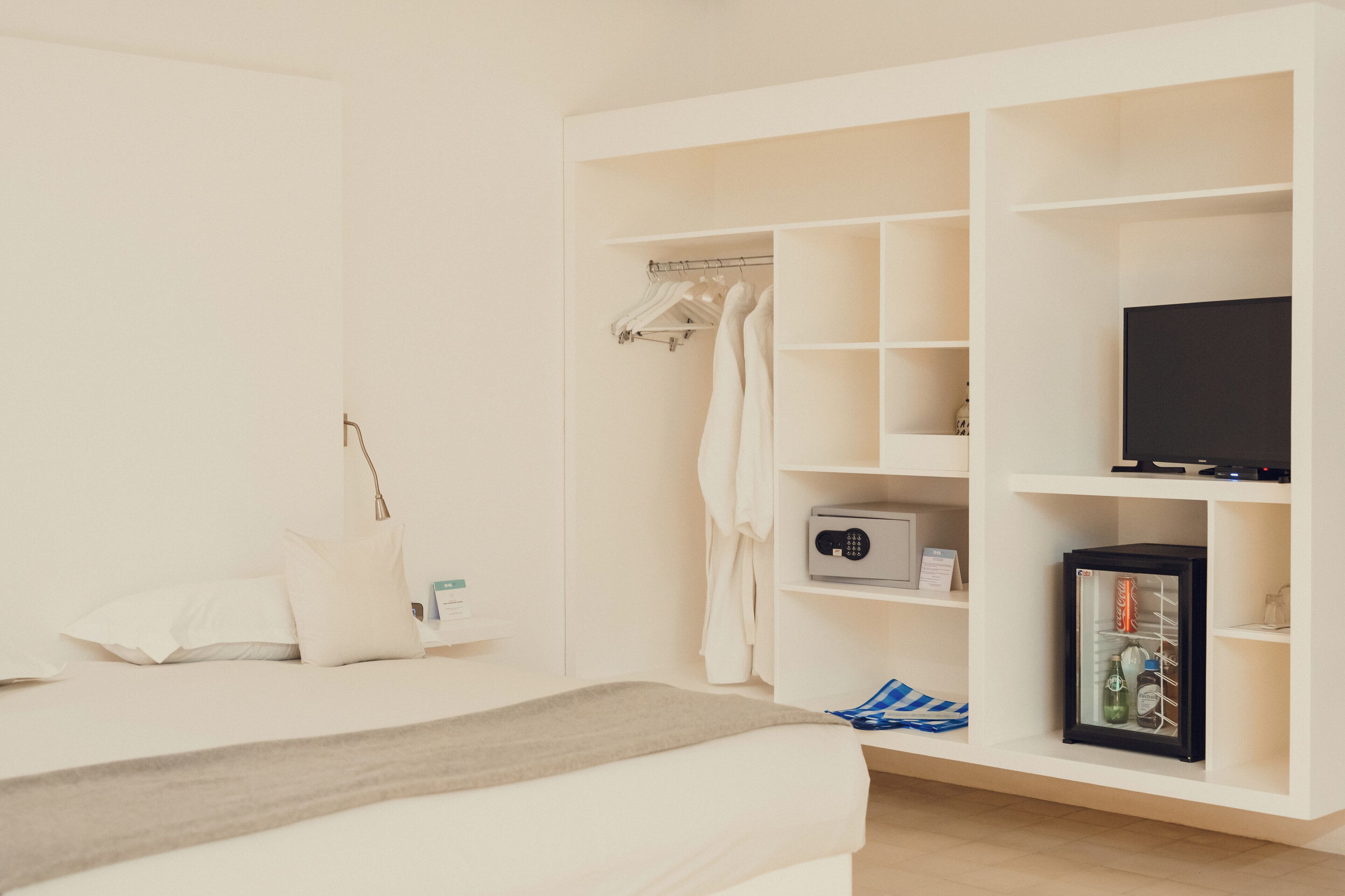 A bedroom with a bed, a wardrobe with hanging clothes, a television, and a minibar.