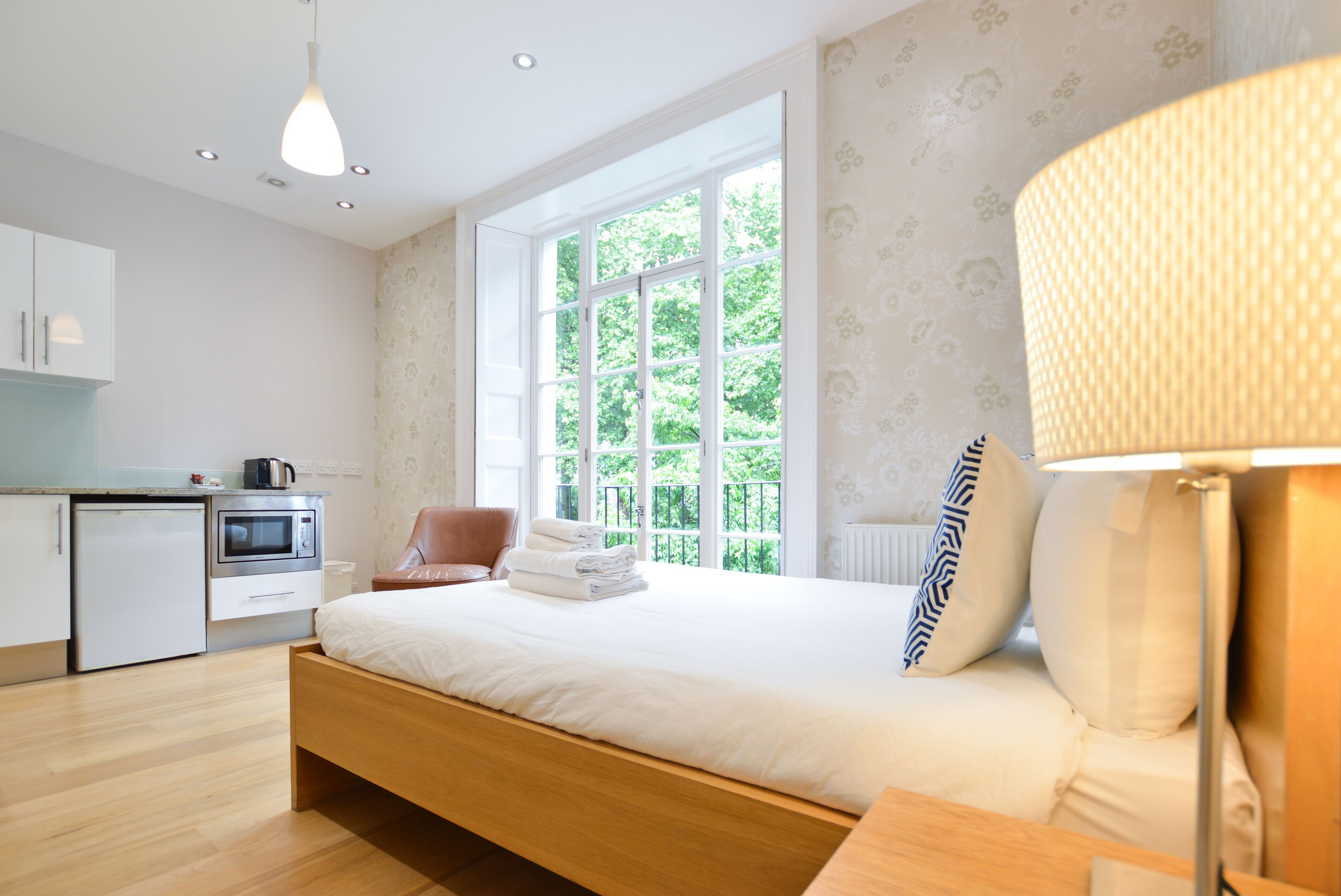 A bedroom with a bed, a bedside table, a lamp, a window with a view of greenery, a kitchenette with a microwave, and a cabinet.