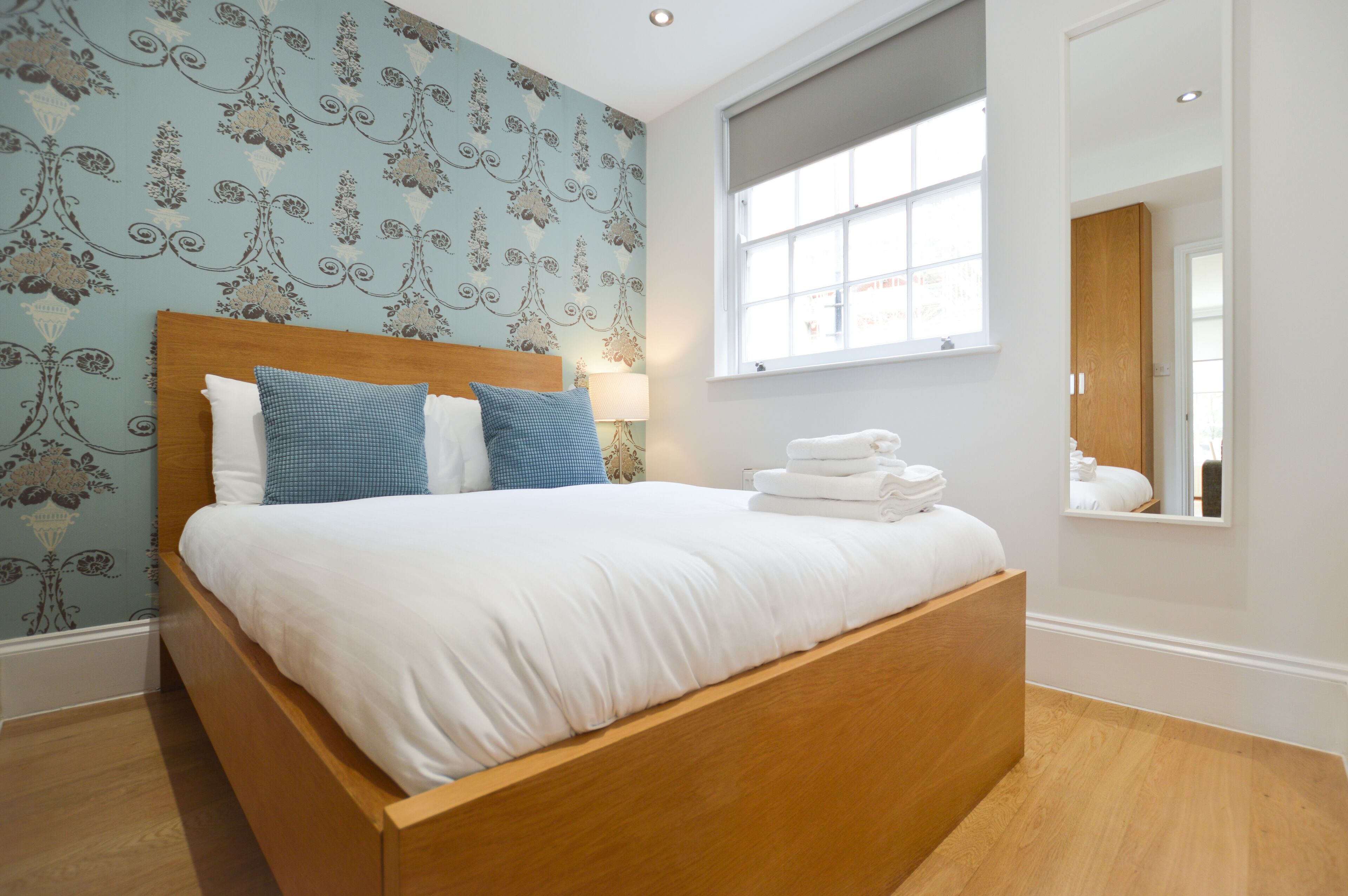 A neatly made bed with white linens and blue pillows, a wooden headboard, and a window with a view of the city.