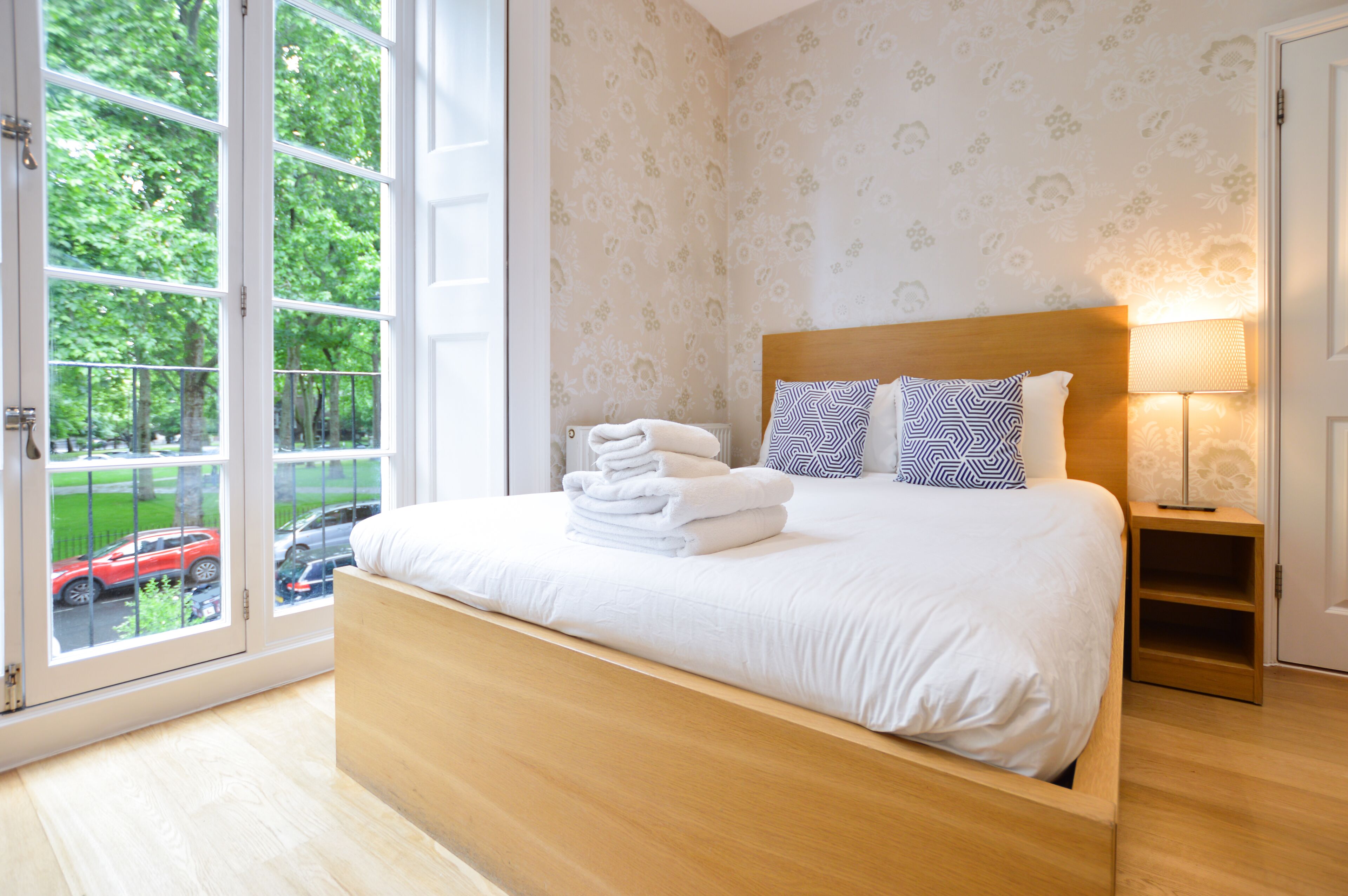 Een slaapkamer met een groot raam, een houten bed en een nachtkastje met een lamp.