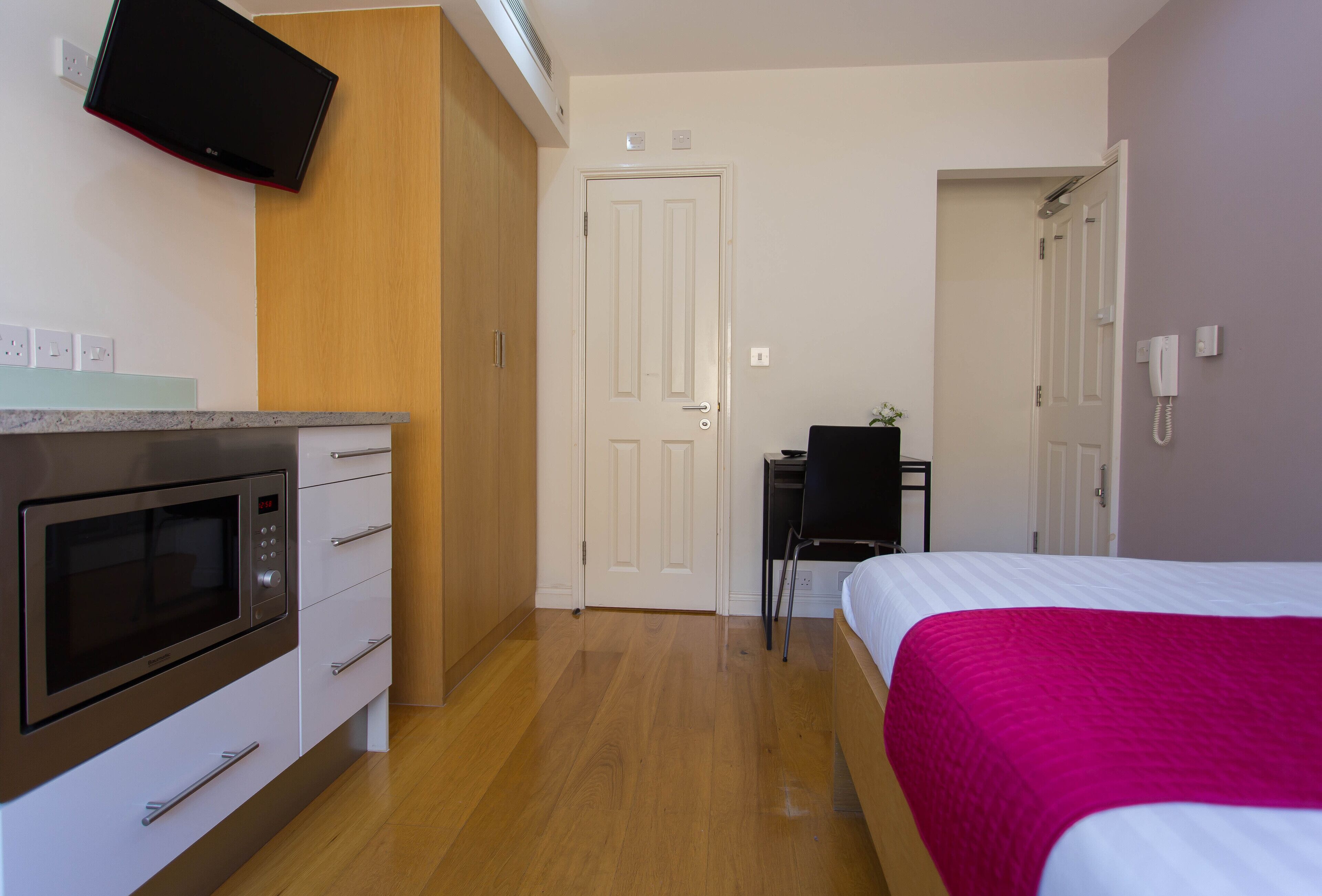 A bedroom with a bed, a desk, a chair, a microwave, and a TV mounted on the wall.