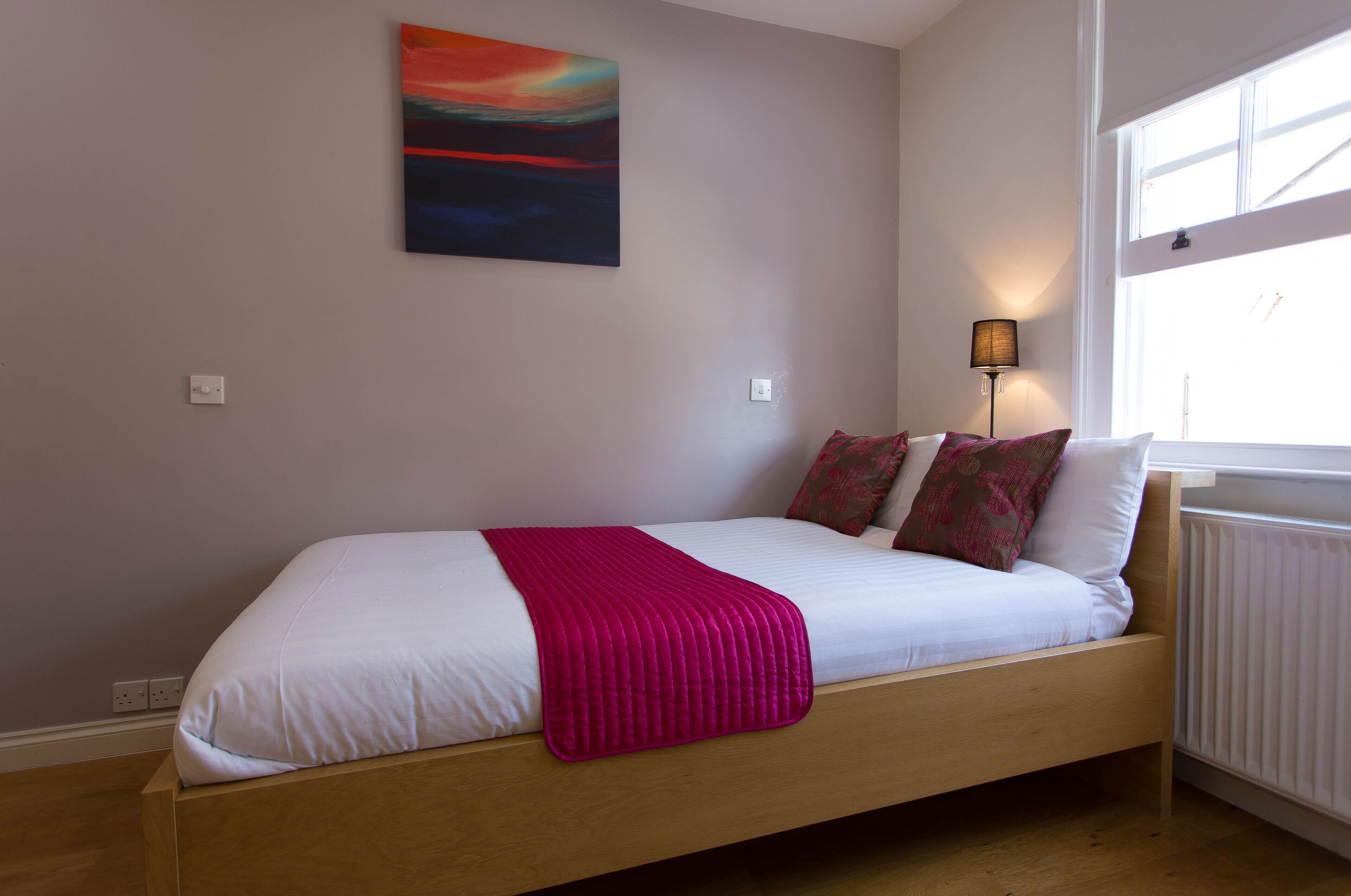 A single bed with a wooden frame, white bedding, and a pink throw blanket. There is a colorful abstract painting on the wall above the bed.