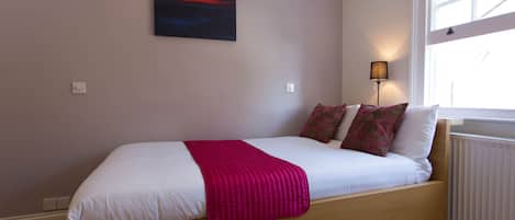 A single bed with a wooden frame, white bedding, and a pink throw blanket. There is a colorful abstract painting on the wall above the bed.