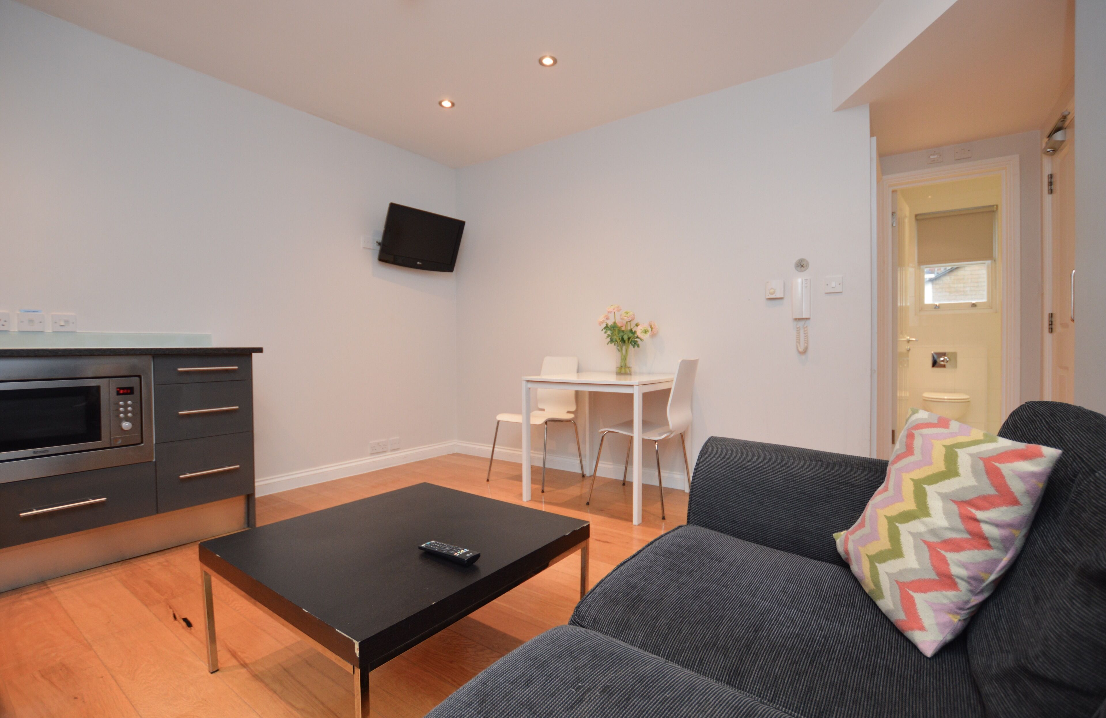 Ein modernes Wohnzimmer mit grauem Sofa, einem schwarzen Couchtisch, einem kleinen weißen Tisch mit Stühlen, einem an der Wand befestigten Flachbildfernseher und einer Küchenzeile mit Mikrowelle und Schränken.