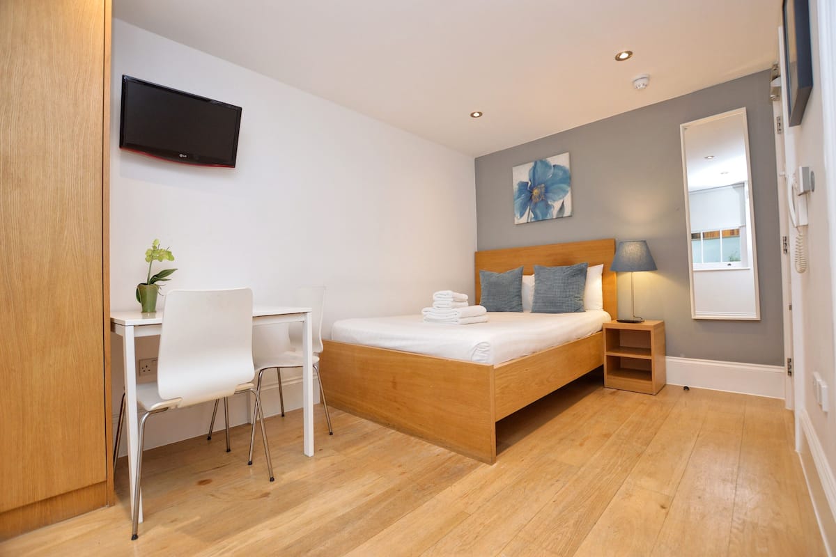 Ein modernes Schlafzimmer mit einem hölzernen Bett, einem Schreibtisch, einem Stuhl, einem an der Wand befestigten Fernseher und einem kleinen Nachttisch mit Lampe.