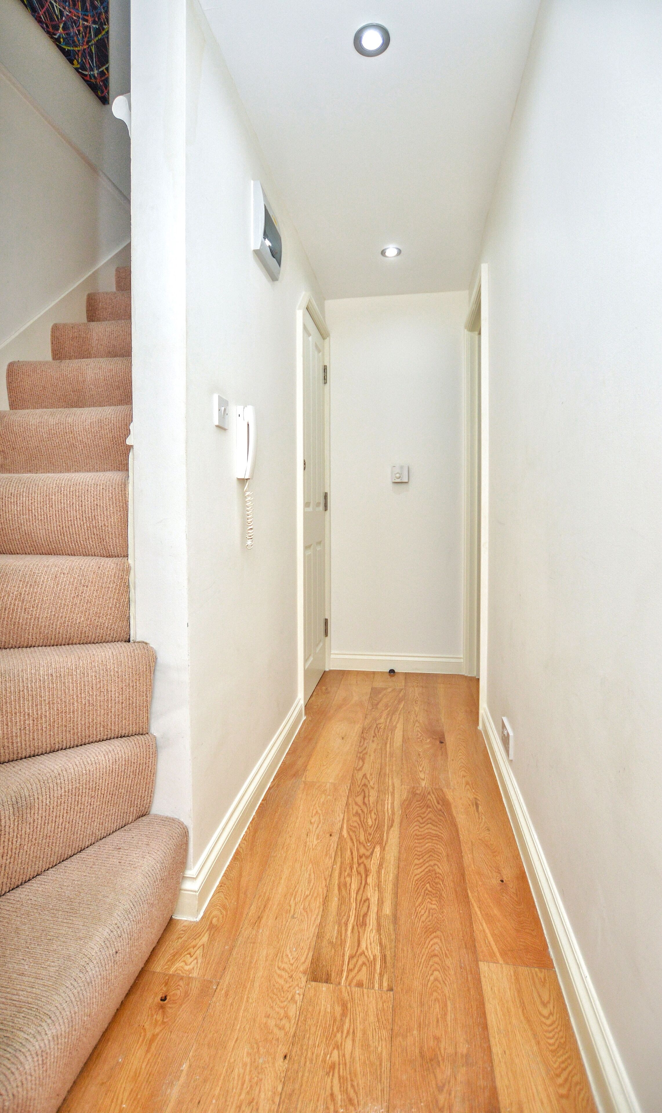 A narrow hallway with wooden flooring, white walls, and a carpeted staircase on the left.