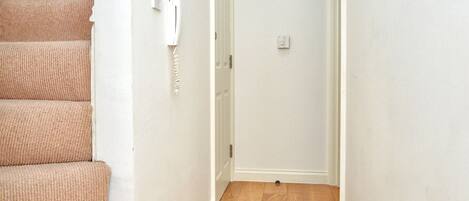 A narrow hallway with wooden flooring, white walls, and a carpeted staircase on the left.