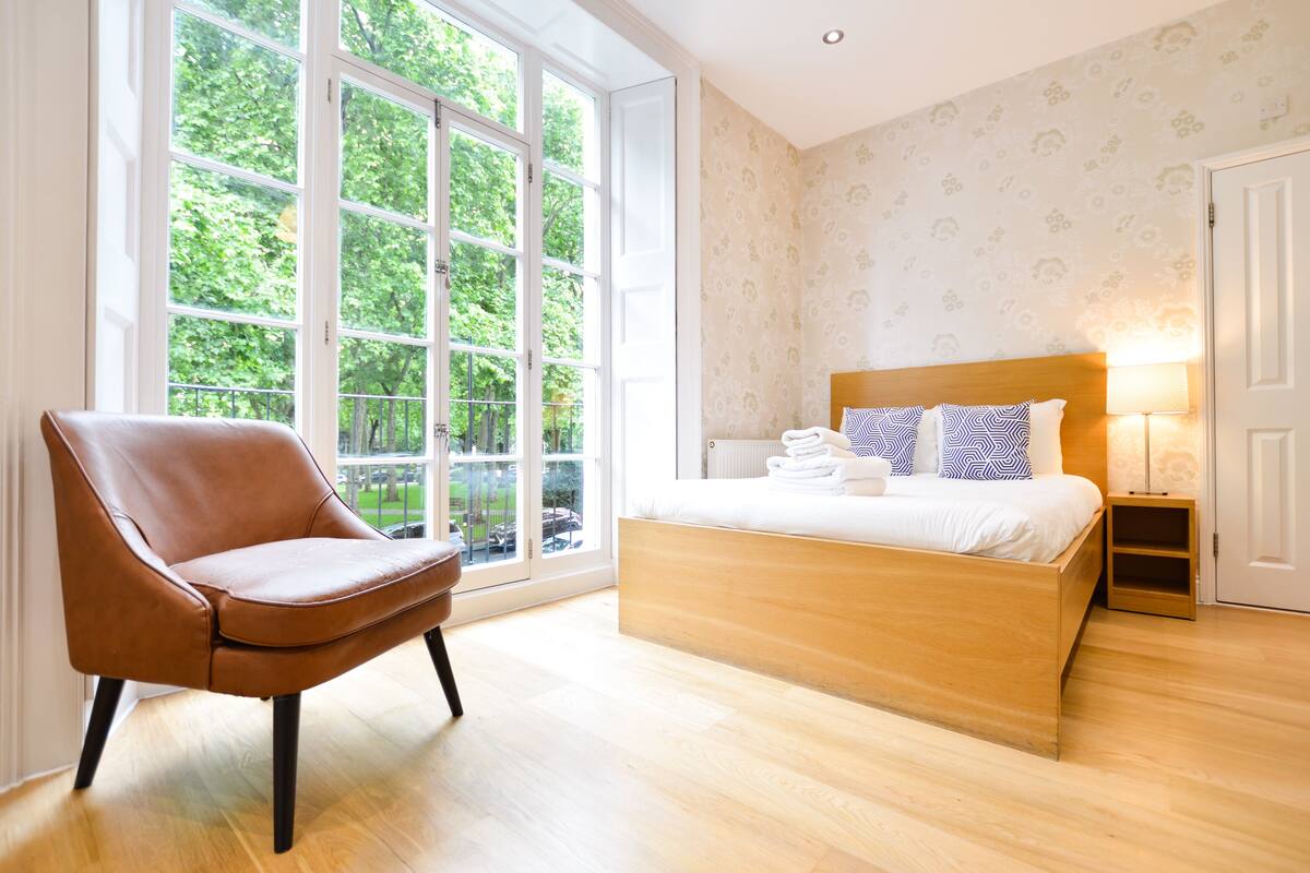 Ein Schlafzimmer mit großem Fenster, einem hölzernen Bett, einem braunen Ledersessel und einem Nachttisch mit Lampe.