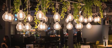 A restaurant with hanging light bulbs and greenery. Tables and chairs are arranged for dining.