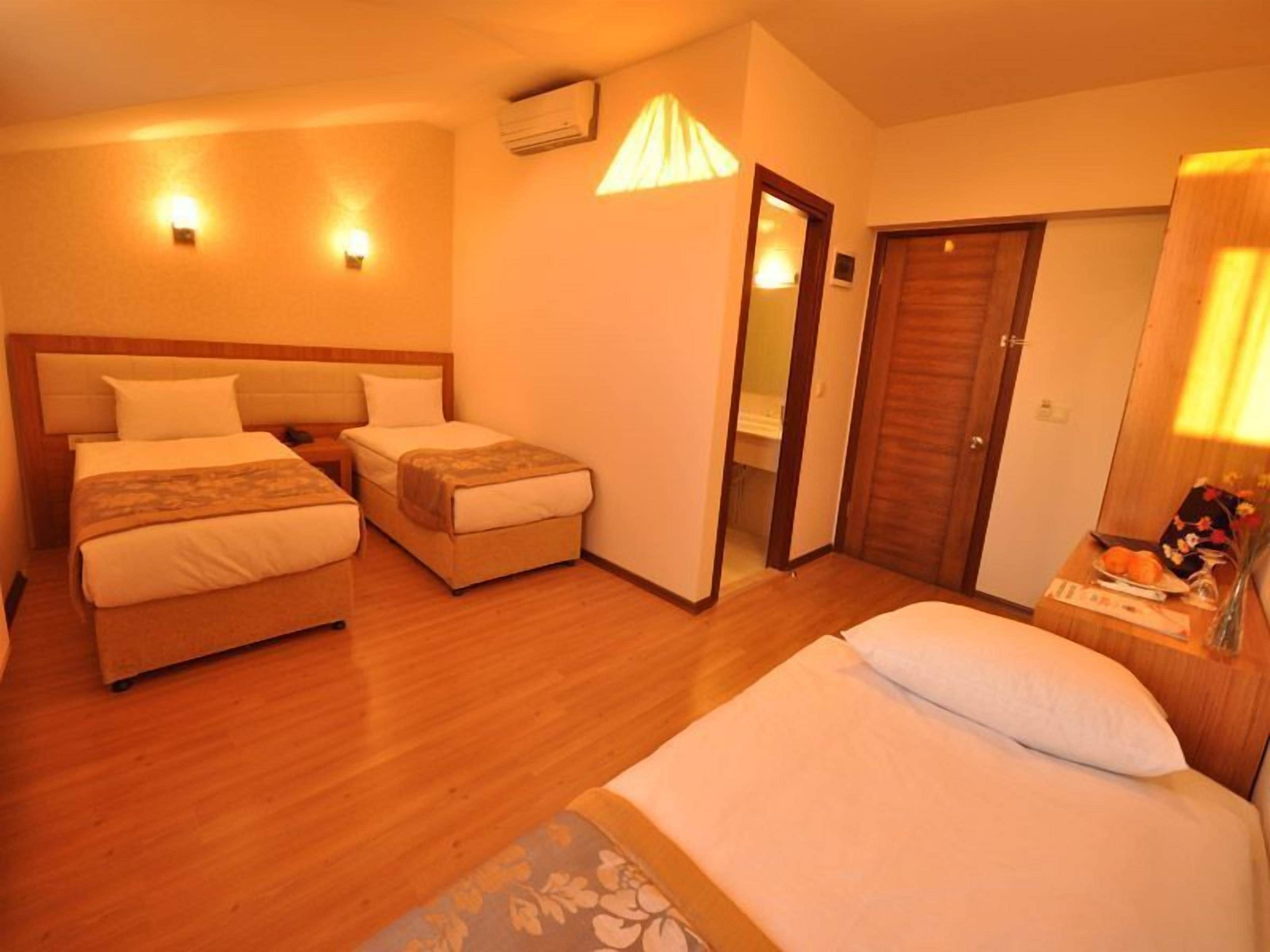 Quarto de hotel com duas camas, piso de madeira, mesa de cabeceira com abajur e porta para o banheiro.
