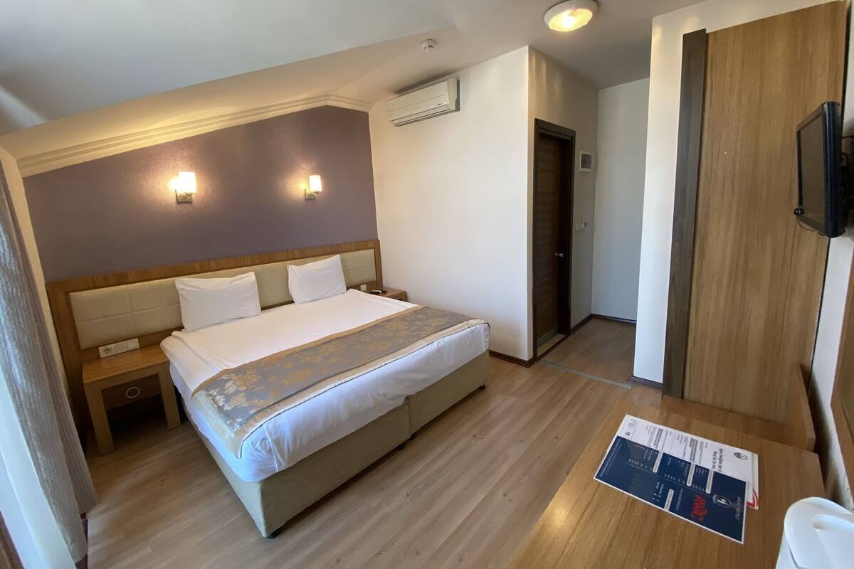 A hotel room with a bed, bedside tables, an air conditioning unit, a wooden door, and a desk with a chair.