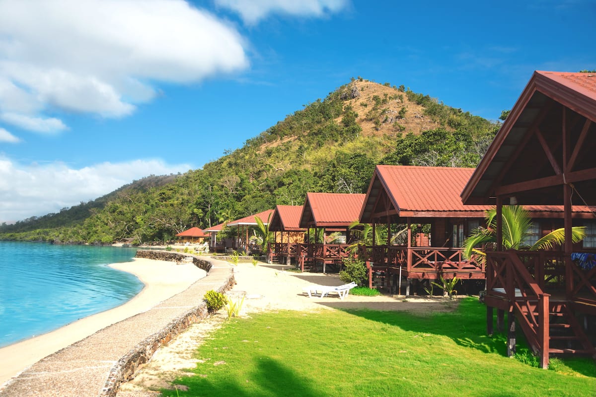 A beachfront resort with wooden cabins, a sandy beach, and a mountain in the background.