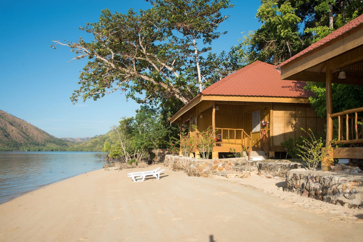 A wooden cabin with a red roof by a lake, surrounded by trees and hills.