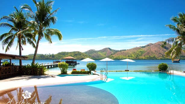 Una piscina de resort con palmeras, una terraza de madera y vistas al agua y las colinas.