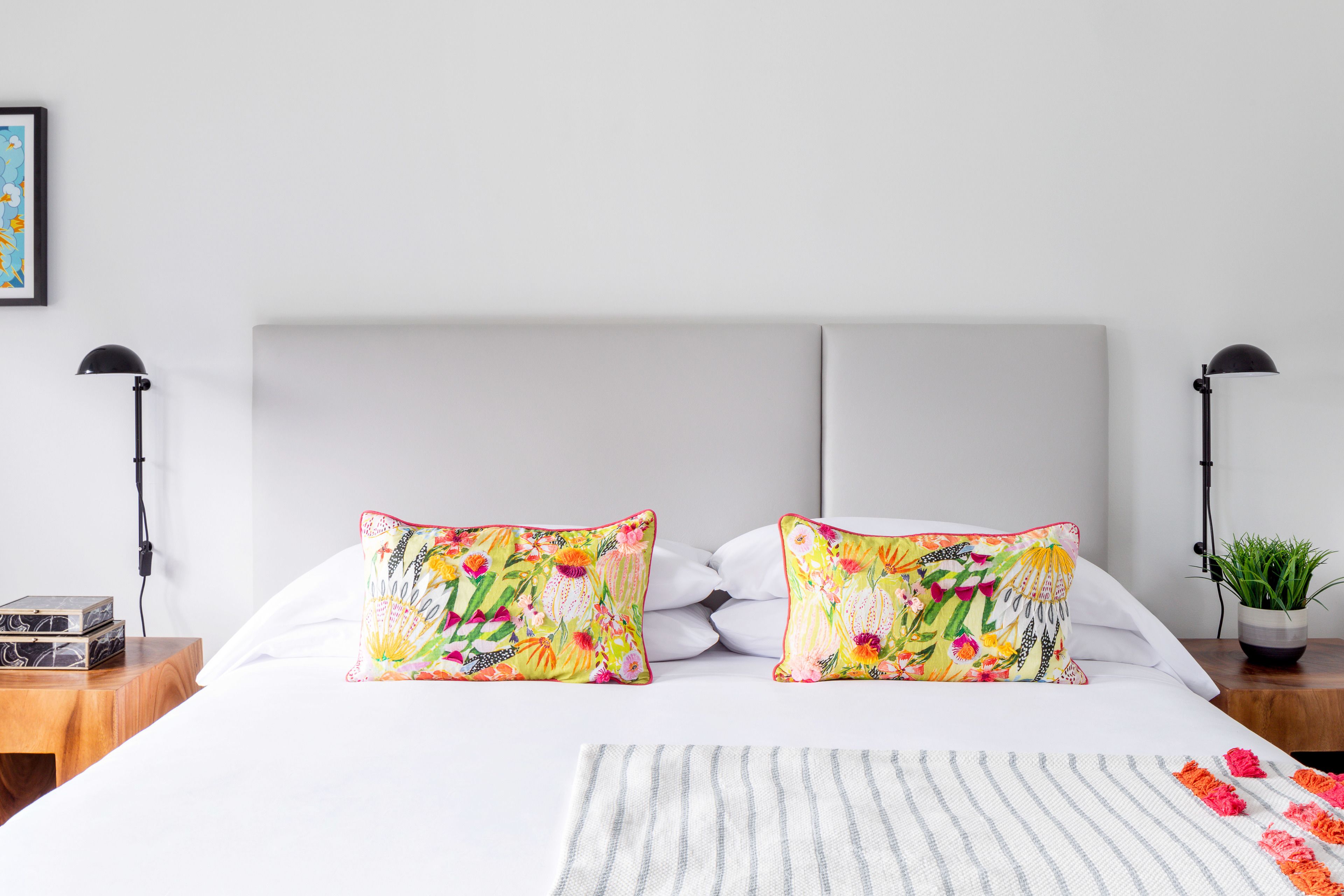Una cama bien hecha con dos almohadas coloridas, una mesita de noche con una lámpara y una pequeña planta en maceta.