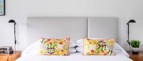 Una cama bien hecha con dos almohadas coloridas, una mesita de noche con una lámpara y una pequeña planta en maceta.