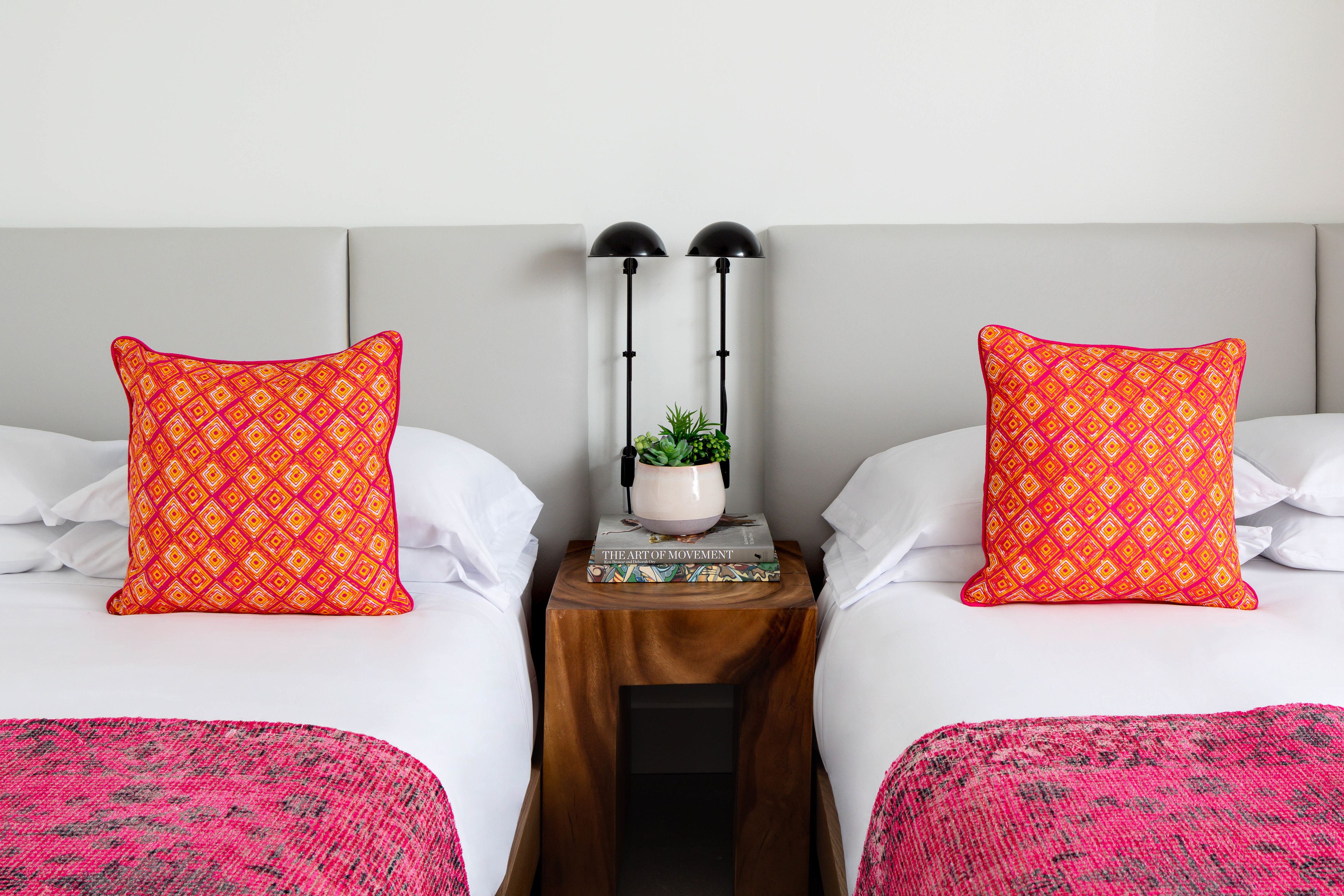 A hotel room with two beds, each with a patterned pillow, a bedside table with a plant, and a book titled "The Art of Movement".