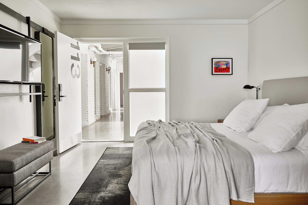 Ein modernes Schlafzimmer mit Bett, Nachttisch und einer Bank. Eine Tür führt zu einem Flur mit einer Treppe.