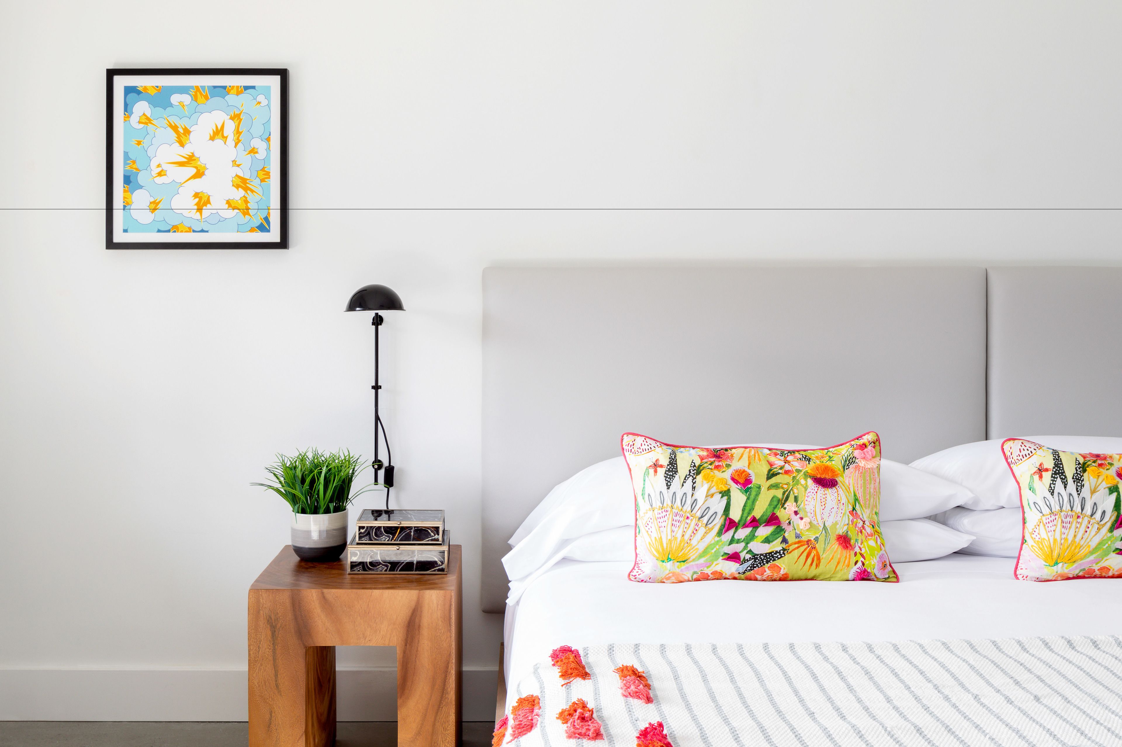 Een slaapkamer met een bed, twee sierkussens met bloemenprint, een nachtkastje met een plant en een lamp, en een lijst met een kunstwerk aan de muur.