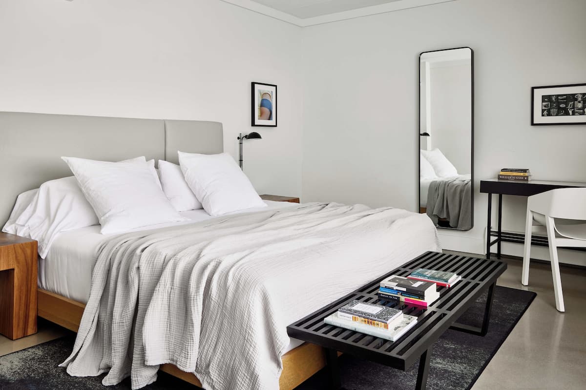 Ein modernes Hotelzimmer mit einem großen Bett, einem Nachttisch, einem Schreibtisch mit Stuhl, einem Spiegel und einem Bücherregal.