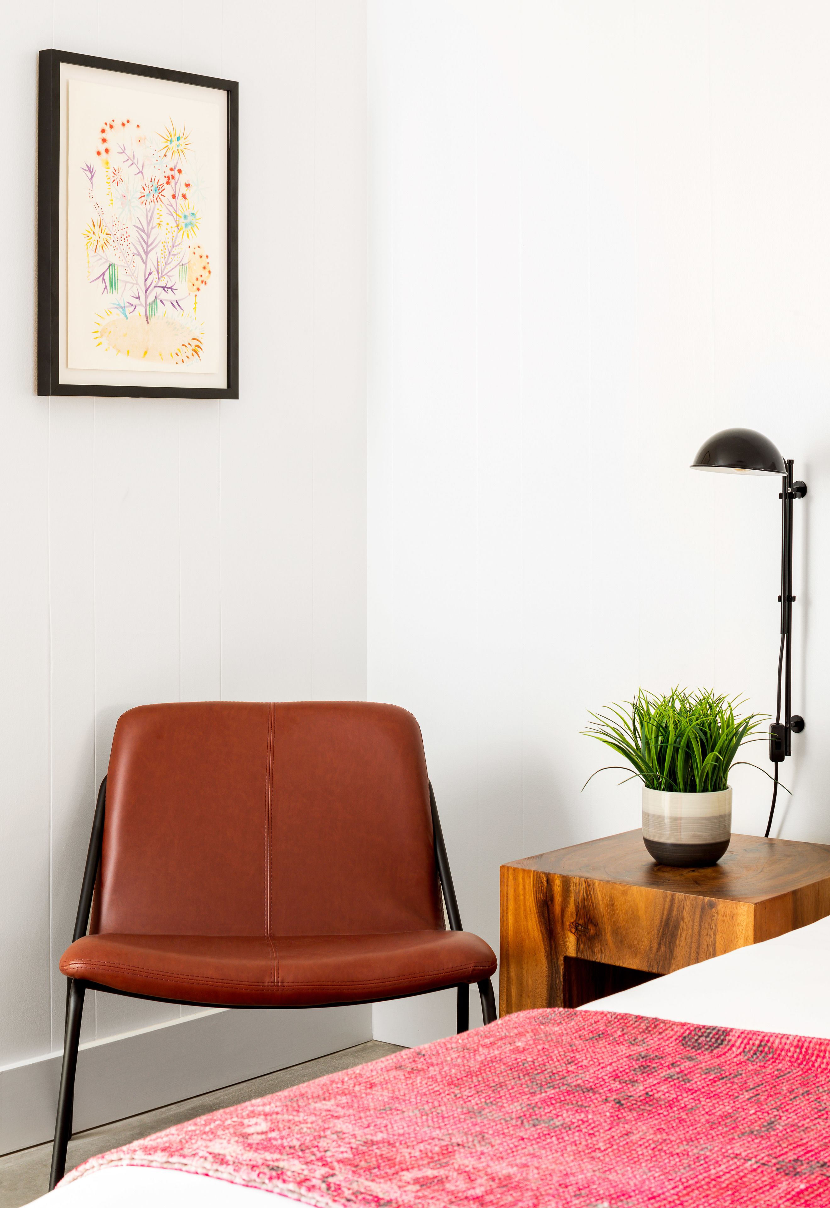 A room with a brown leather chair, a wooden side table with a potted plant, a black floor lamp, and a framed artwork on the wall.