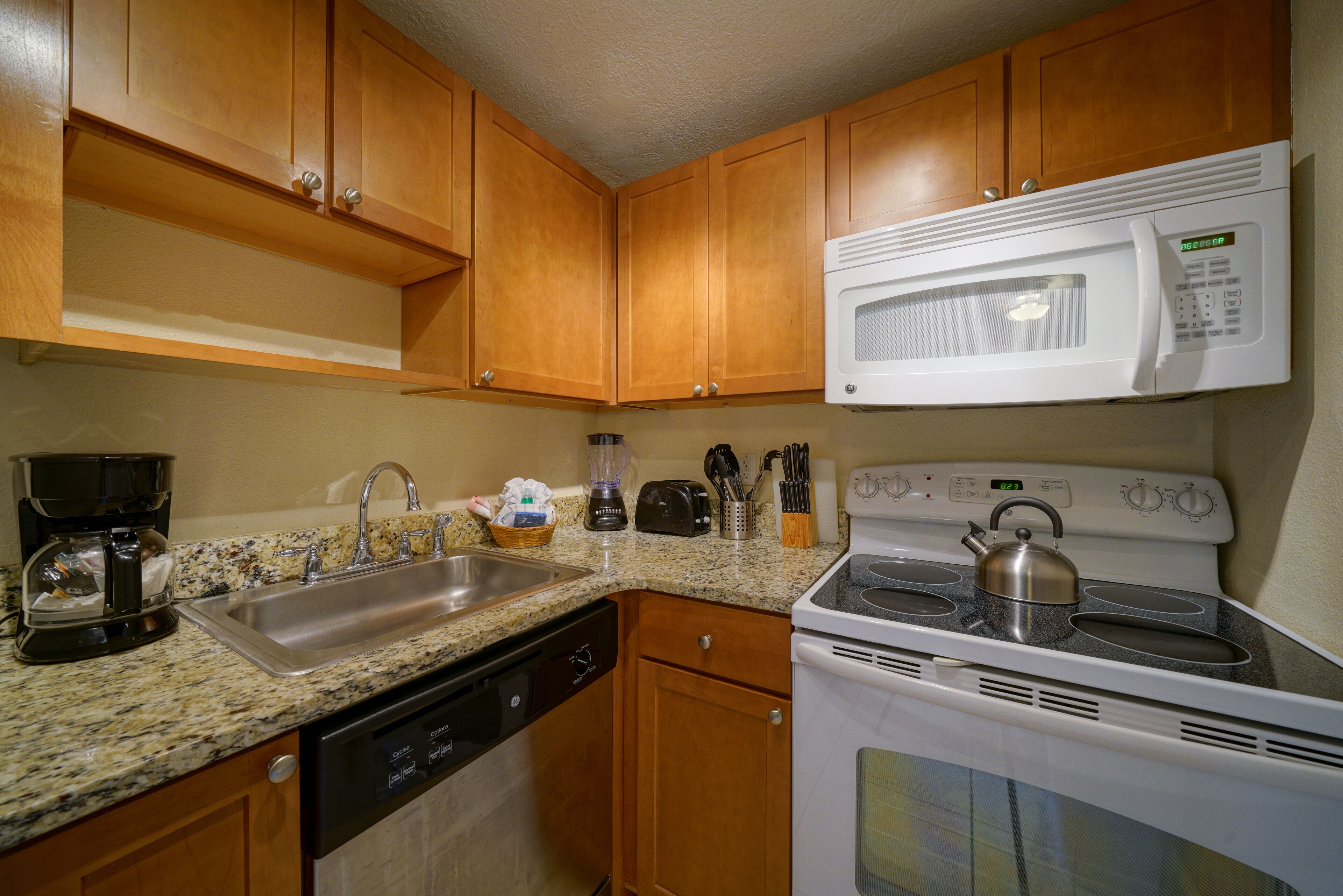 A kitchen with wooden cabinets, a stainless steel sink, a built-in microwave, a stove with a kettle, and a coffee maker.
