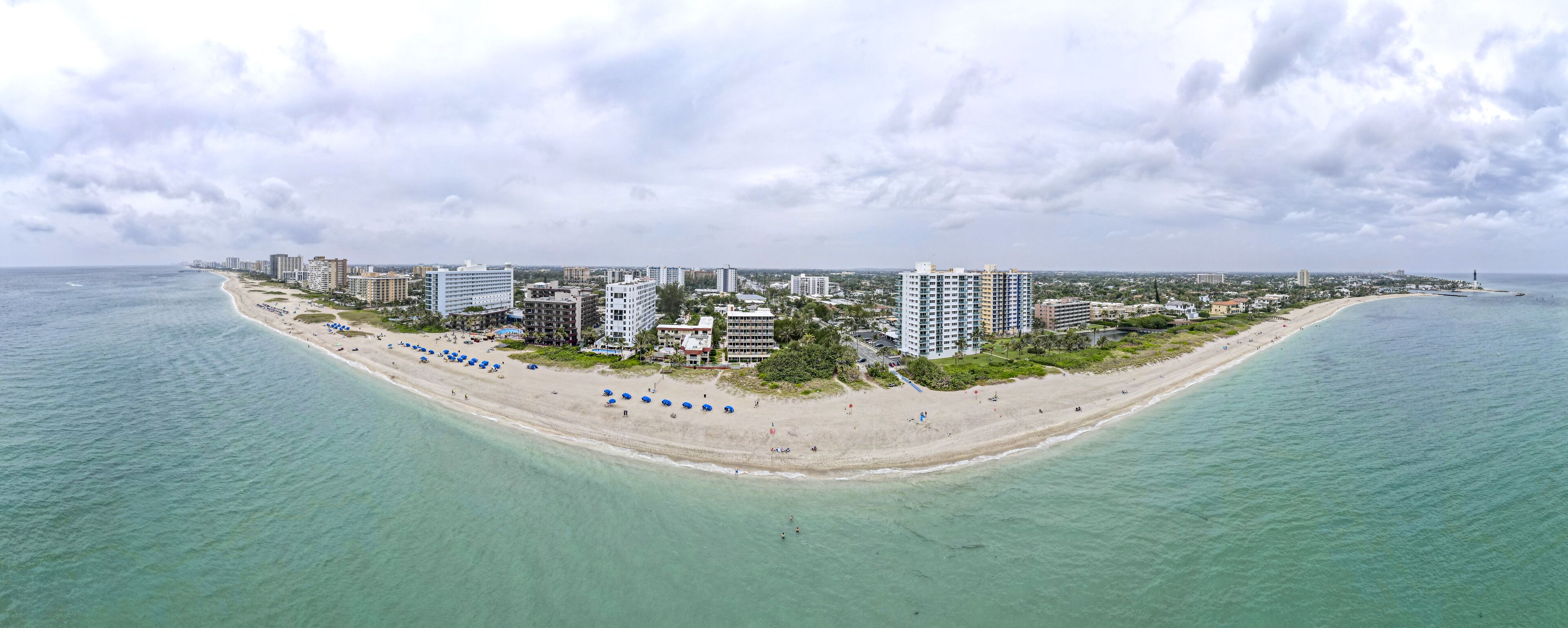 An aerial view of a coastal city with a long sandy beach, numerous high-rise buildings, and a clear view of the ocean.