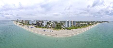 An aerial view of a coastal city with a long sandy beach, numerous high-rise buildings, and a clear view of the ocean.