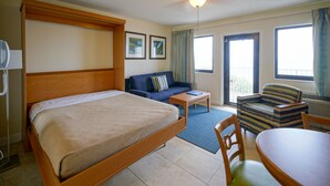 A hotel room with a bed, a sofa, a small table, and a chair. There are two windows and a door. - La Costa Beach Club (Pompano Beach)