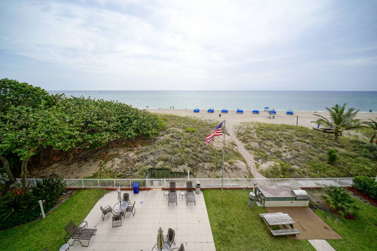 Een terras met tuinmeubilair, een vlag en uitzicht op het strand met parasols.