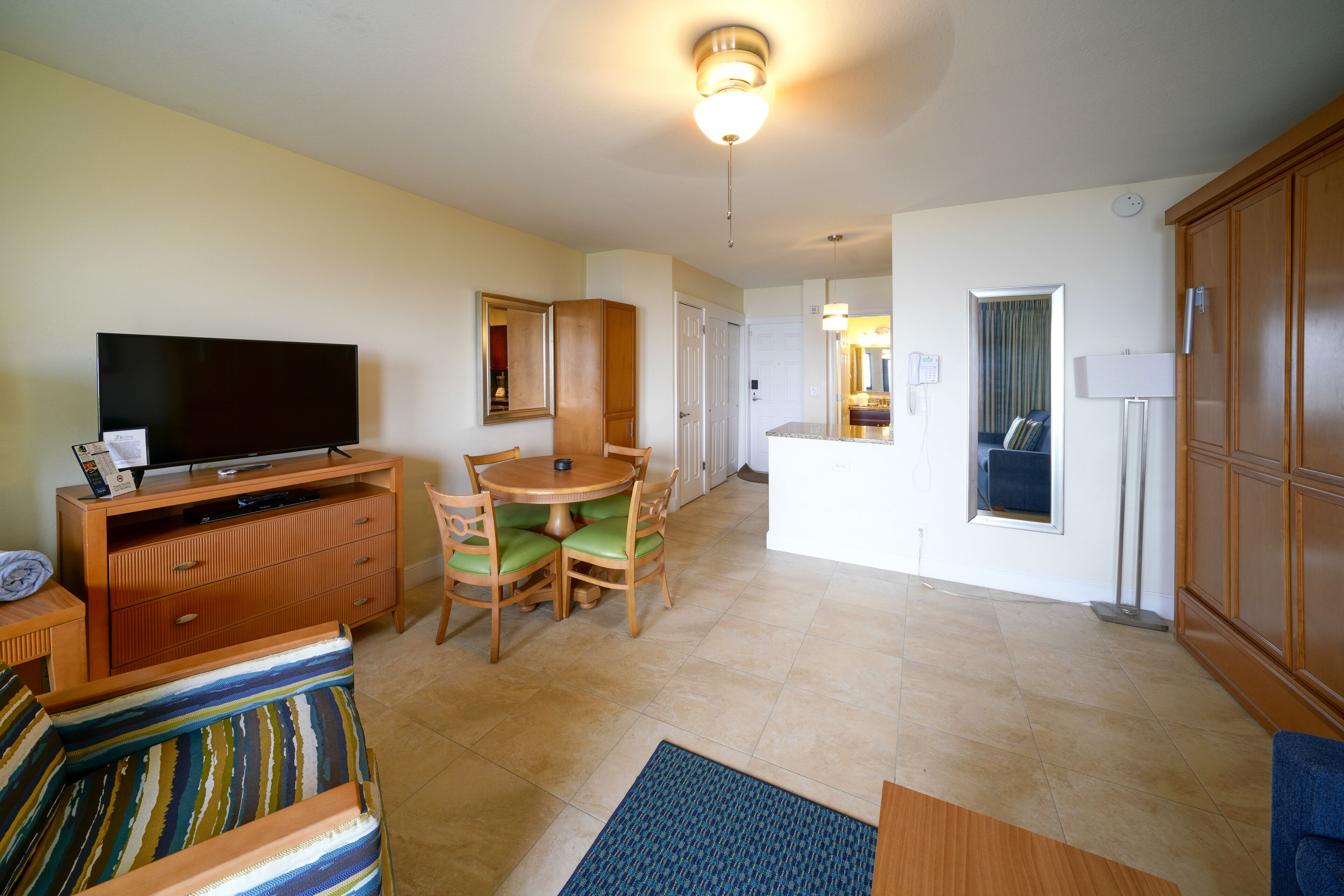 A hotel room with a flat-screen TV, wooden furniture, a dining area, and a bathroom visible through an open door.