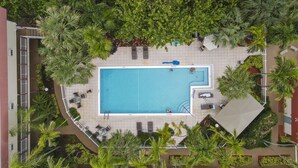 An aerial view of a rectangular swimming pool surrounded by palm trees and lounge chairs, with a tiled deck and a white fence. - La Costa Beach Club (Pompano Beach)