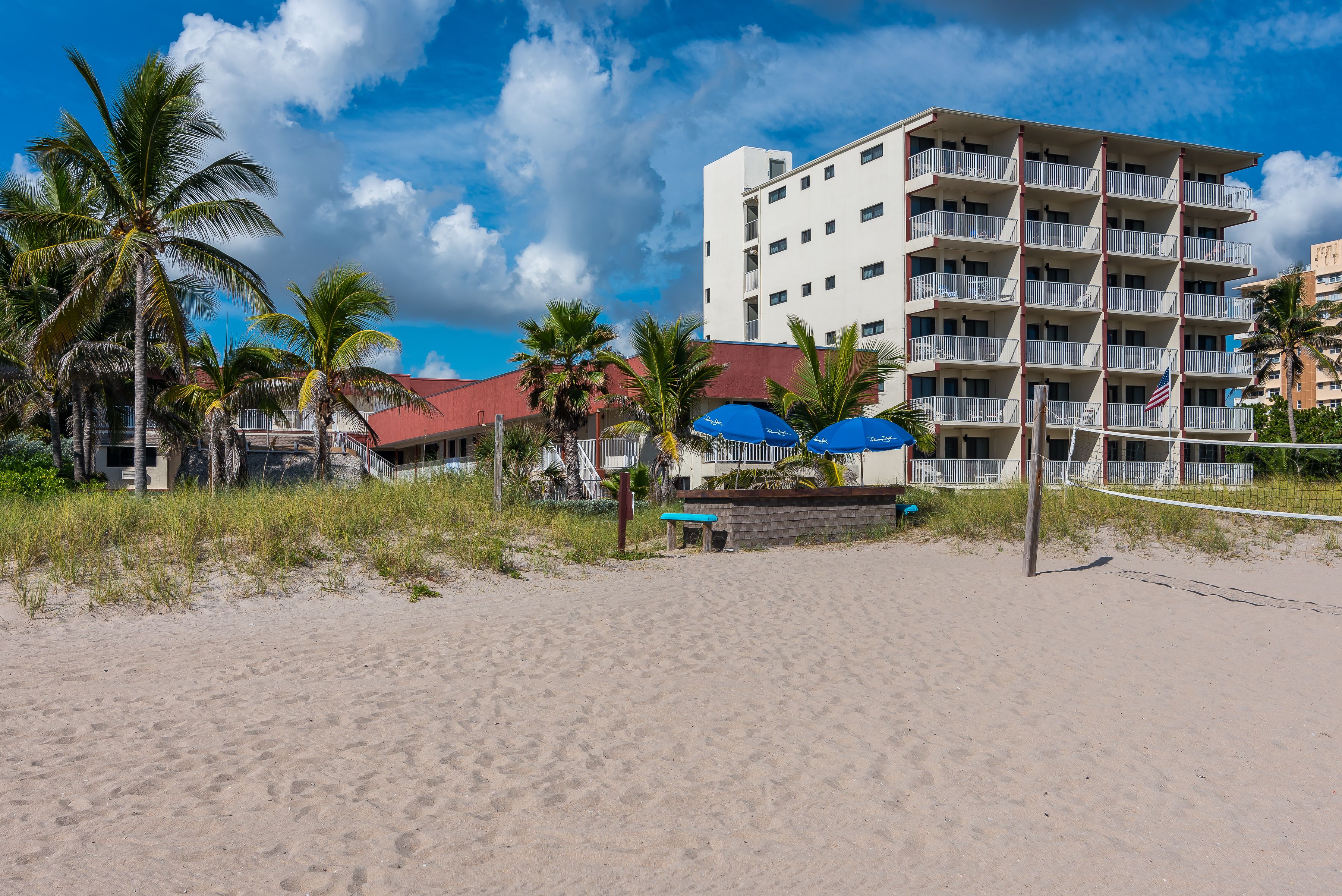 Un hotel fronte spiaggia con numerosi balconi, palme e una rete da pallavolo.