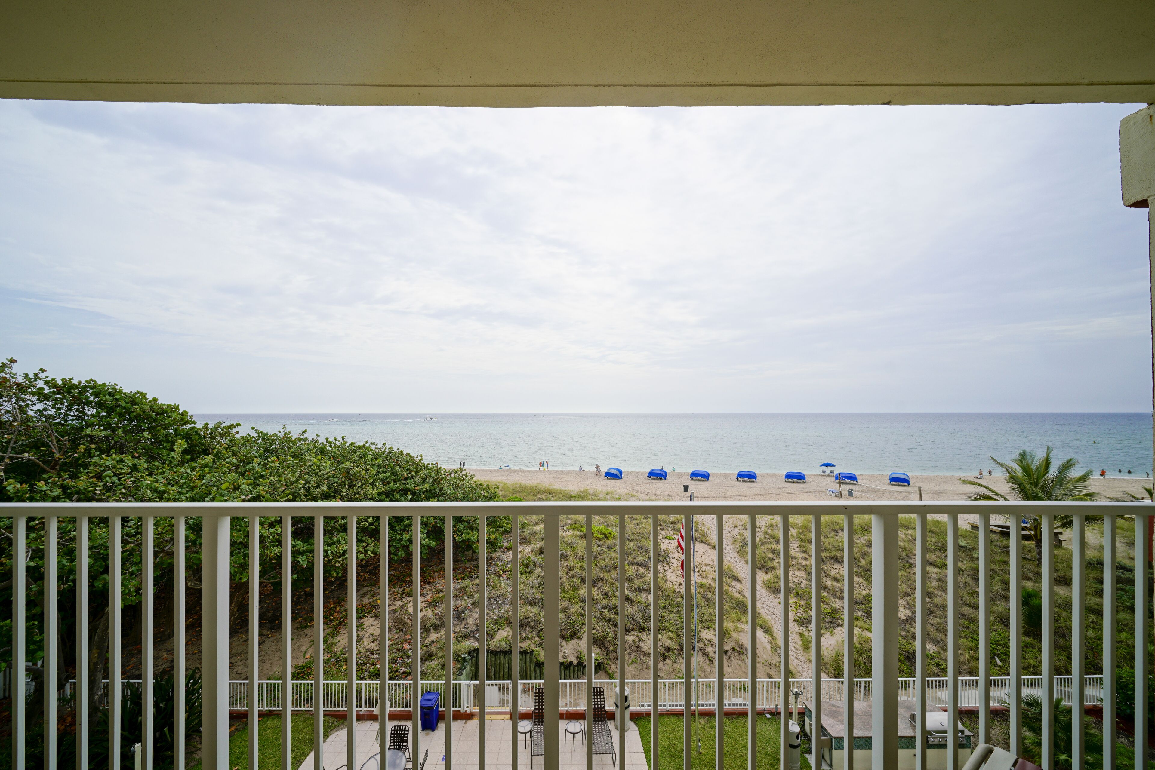 A balcony with a view of the beach, including a pathway, chairs, and umbrellas.
