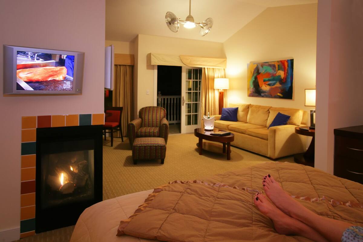 Hotellihuone, jossa on takka, televisio, sohva, nojatuoli ja taulu seinällä.