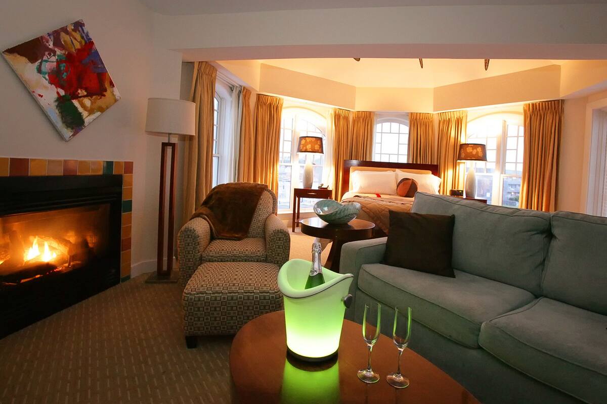 Hotellihuone, jossa on takka, vihreä jäähdytysastia, kaksi lasia, sohva, kuviollinen nojatuoli, sivupöytä lampun kanssa ja taulu seinällä.